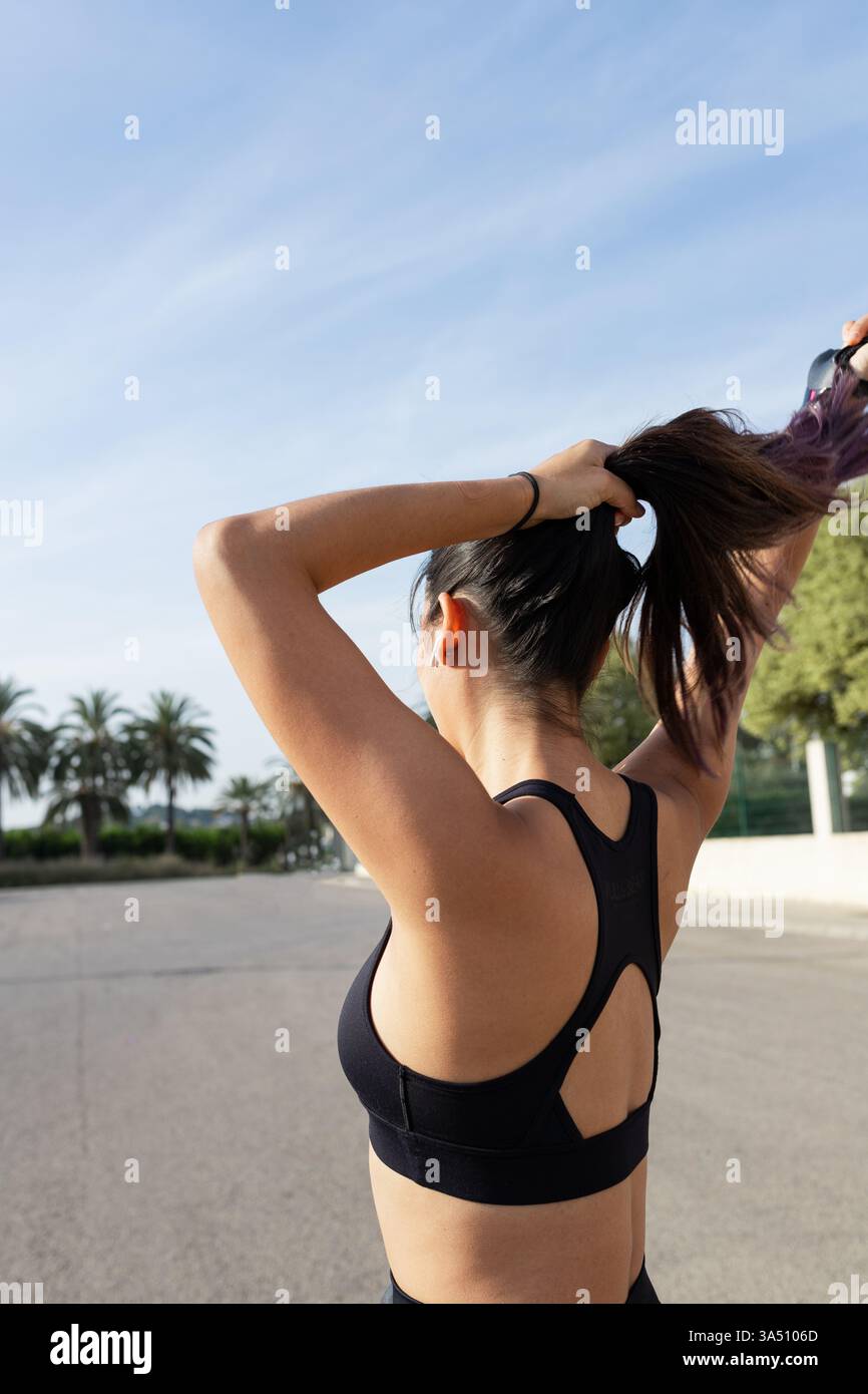 Athlète hispanique féminine en plein air par une journée ensoleillée, se nouant les cheveux avant un entraînement. Elle porte des vêtements de fitness et des écouteurs sans fil pour un style de vie actif moderne. Ce portrait lumineux de fitness convient aux campagnes athlétiques, de bien-être et de vêtements de sport. Banque D'Images