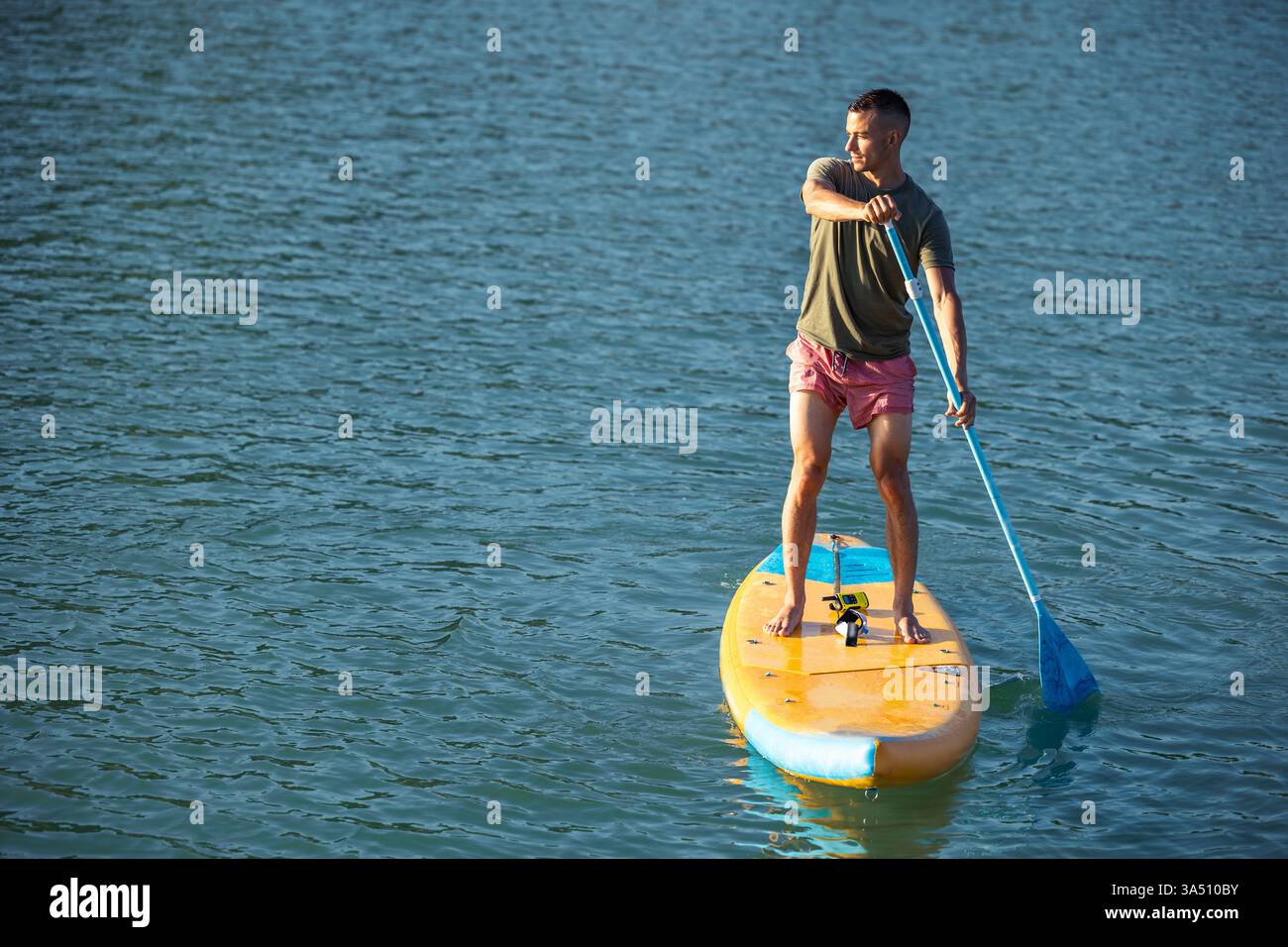 Homme hispanique pagaie sur une planche à pagaie à la plage, regardant loin en été. Une image dynamique d'activité côtière mettant en valeur le stand up paddleboard et les loisirs en bord de mer. Idéal pour les campagnes marketing de voyage, de sport et de vacances. Banque D'Images