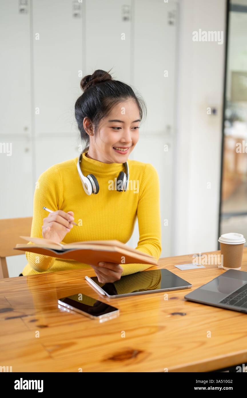 Une étudiante universitaire asiatique étudie à l'intérieur, recherchant des informations en ligne et dans un manuel à son bureau. Ce portrait éducatif correspond à la vie universitaire et aux contextes d’études, véhiculant concentration et ambition académique. Banque D'Images