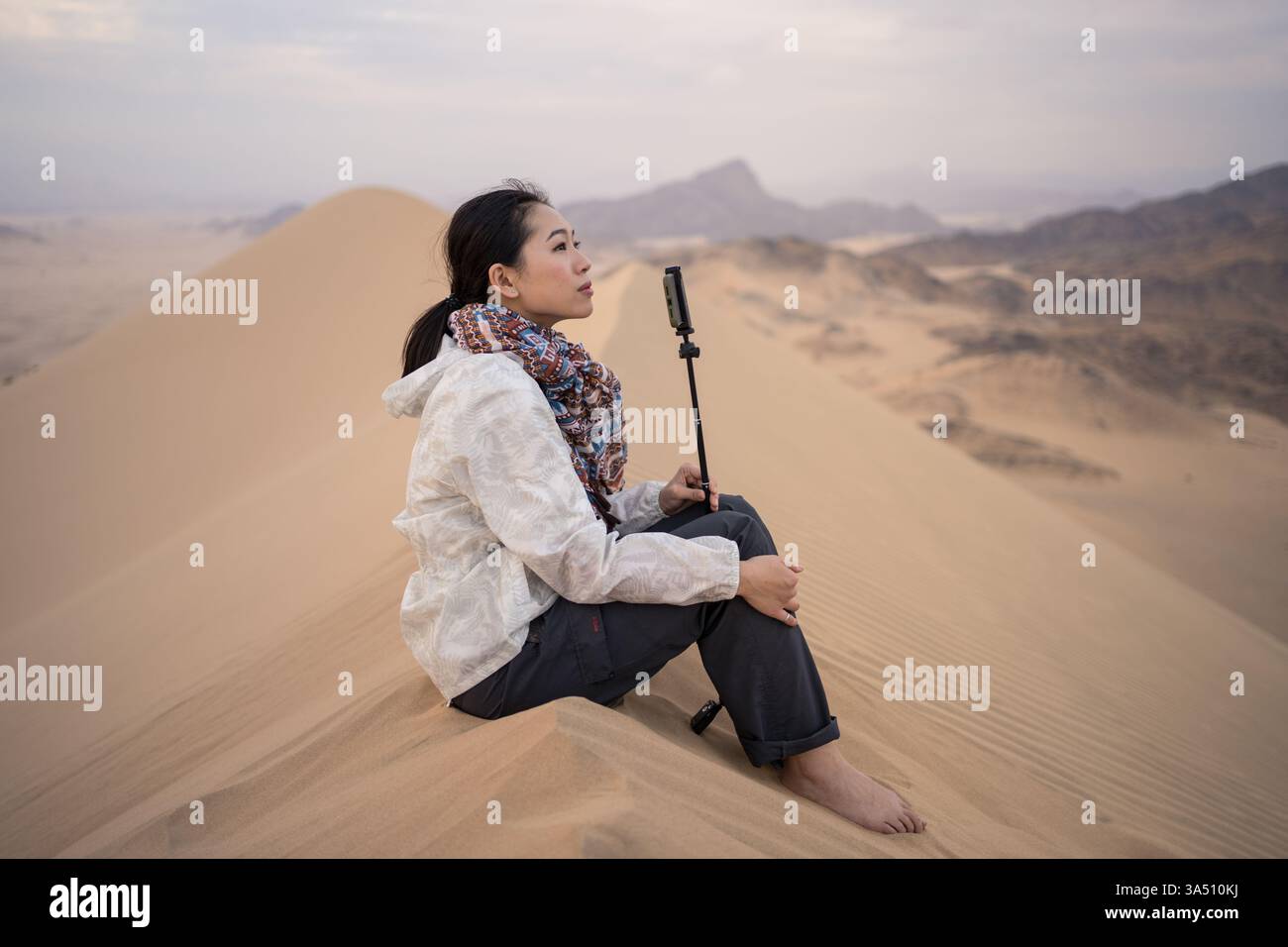 Femme asiatique contemplant un paysage désertique en Arabie Saoudite, assise sur une dune de sable et regardant loin. Cette scène de voyage tranquille met en valeur les paysages naturels, la solitude et l'inspiration. Idéal pour les voyages, le tourisme et les campagnes lifestyle. Banque D'Images