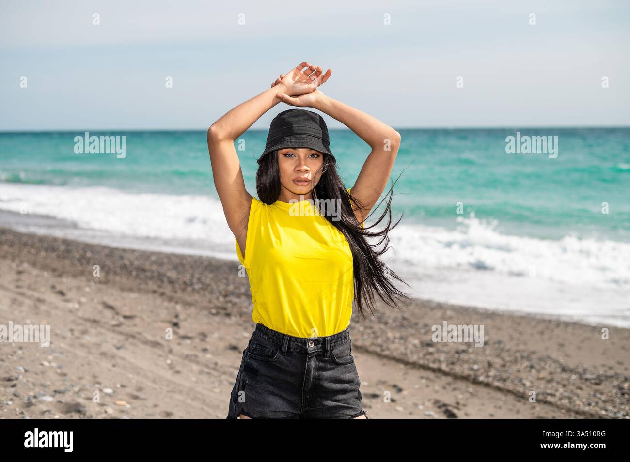 Sérieuse femme afro-américaine debout sur une plage ensoleillée portant un chapeau seau. Elle regarde vers l'avant avec une expression ciblée, mêlant mode et vibrations de voyage. Idéal pour le style de vie, les voyages et les campagnes estivales définies par la côte. Banque D'Images