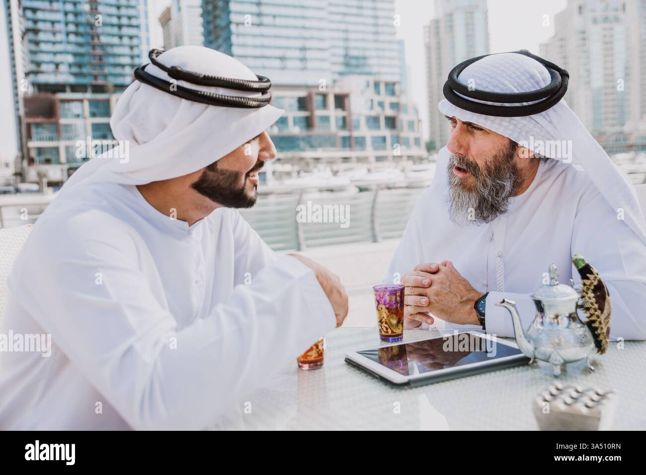 Souriants hommes d'affaires du moyen-Orient et du Caucase portant des kanduras assis dans un café en plein air partageant le thé. Ce moment d’affaires interculturel est adapté à la collaboration internationale et aux thèmes de l’hospitalité. Banque D'Images