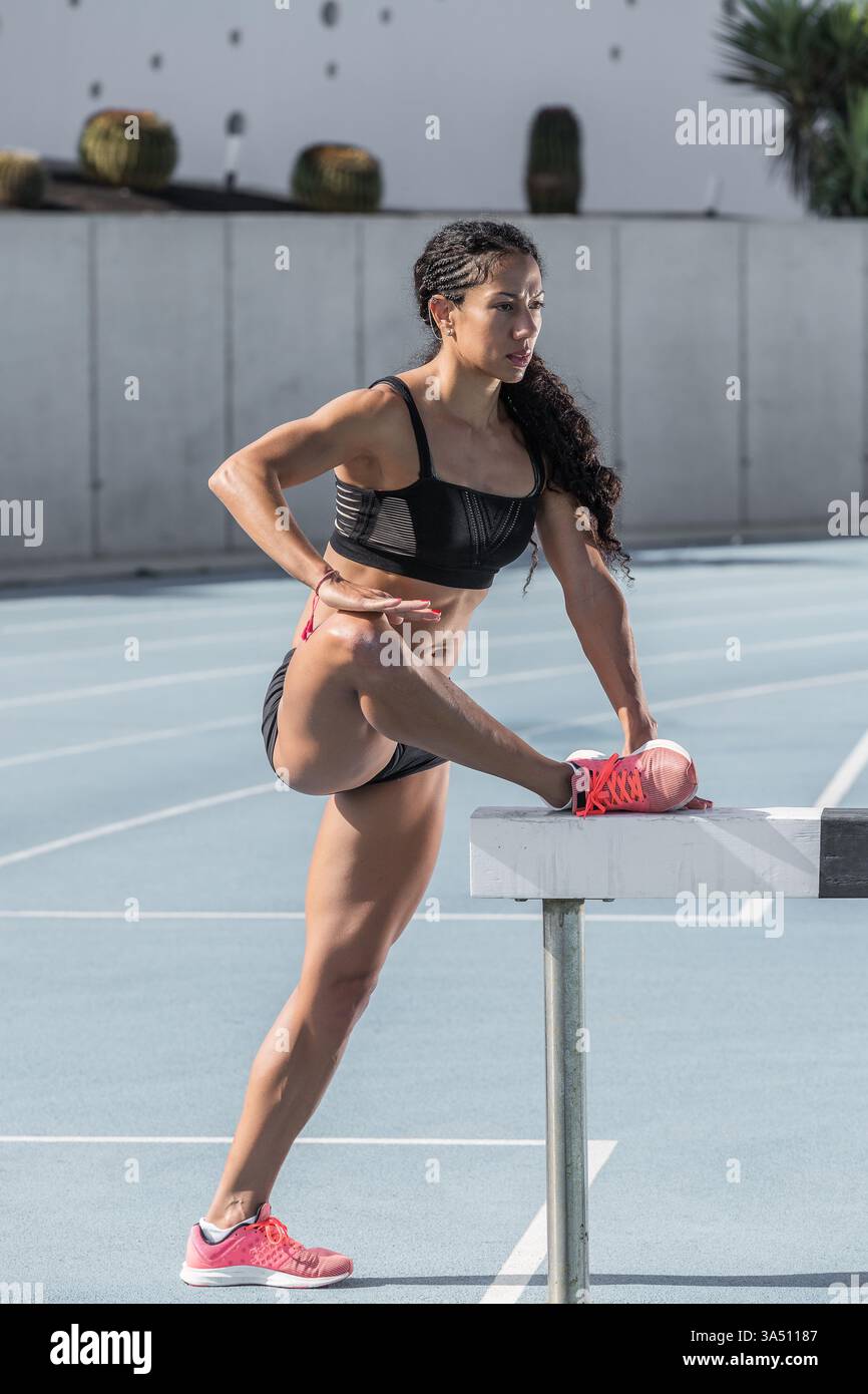 Femme hispanique en vêtements de sport effectue un étirement de jambe sur une piste de course, capturé lors d'un entraînement extérieur frais. Cette image sportive dynamique est idéale pour les campagnes de fitness et les histoires d'athlétisme, mettant en évidence l'échauffement et la préparation sportive. Parfait pour promouvoir les vêtements de sport, l'entraînement en plein air et les styles de vie actifs. Banque D'Images