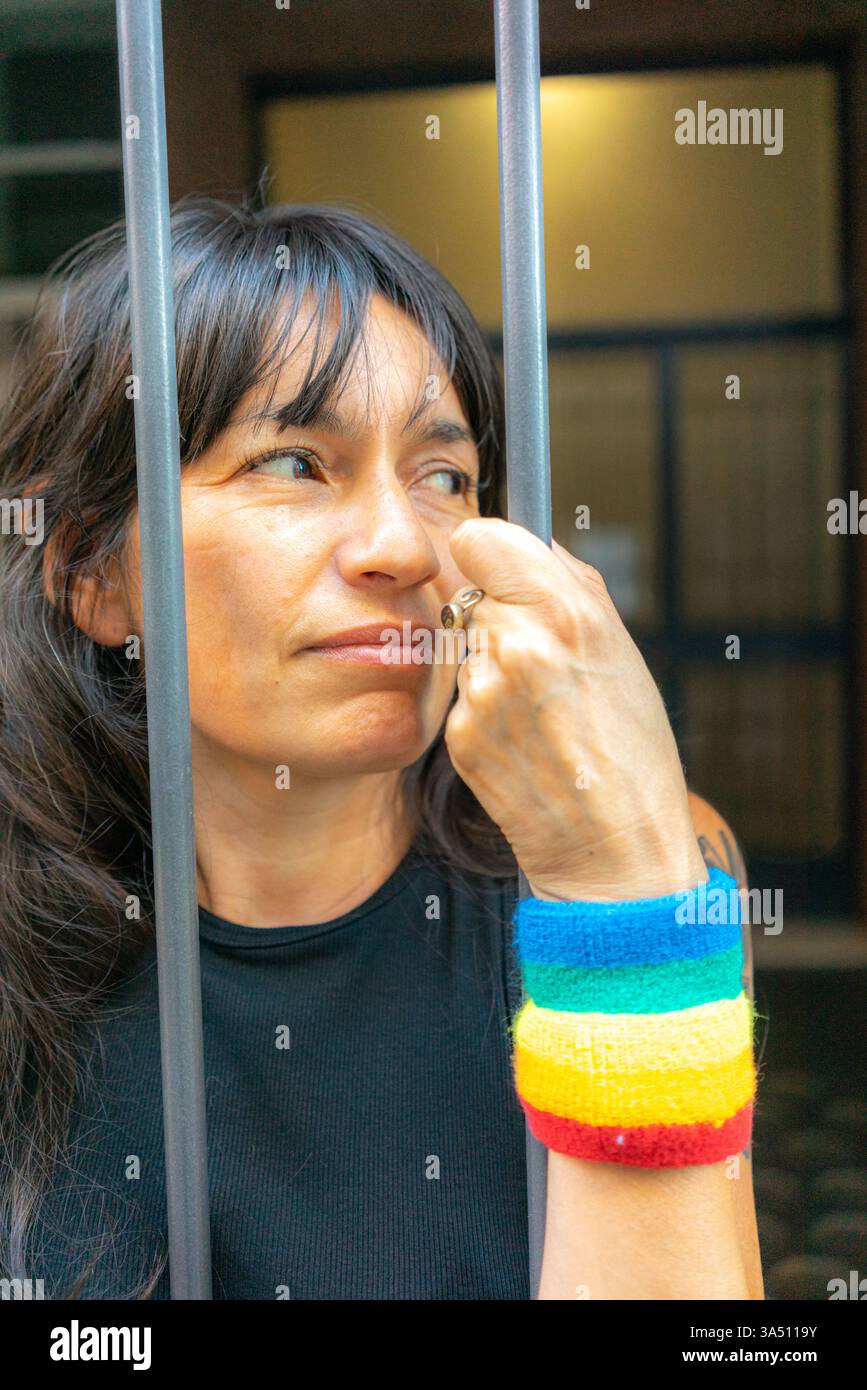 Portrait d'une femme hispanique avec un bracelet arc-en-ciel, debout derrière des balustrades métalliques à la maison et regardant de côté. L'accessoire arc-en-ciel signale la fierté LGBTQ+ et le style personnel. Cette image fonctionne bien pour le style de vie, la diversité et les campagnes communautaires. Banque D'Images