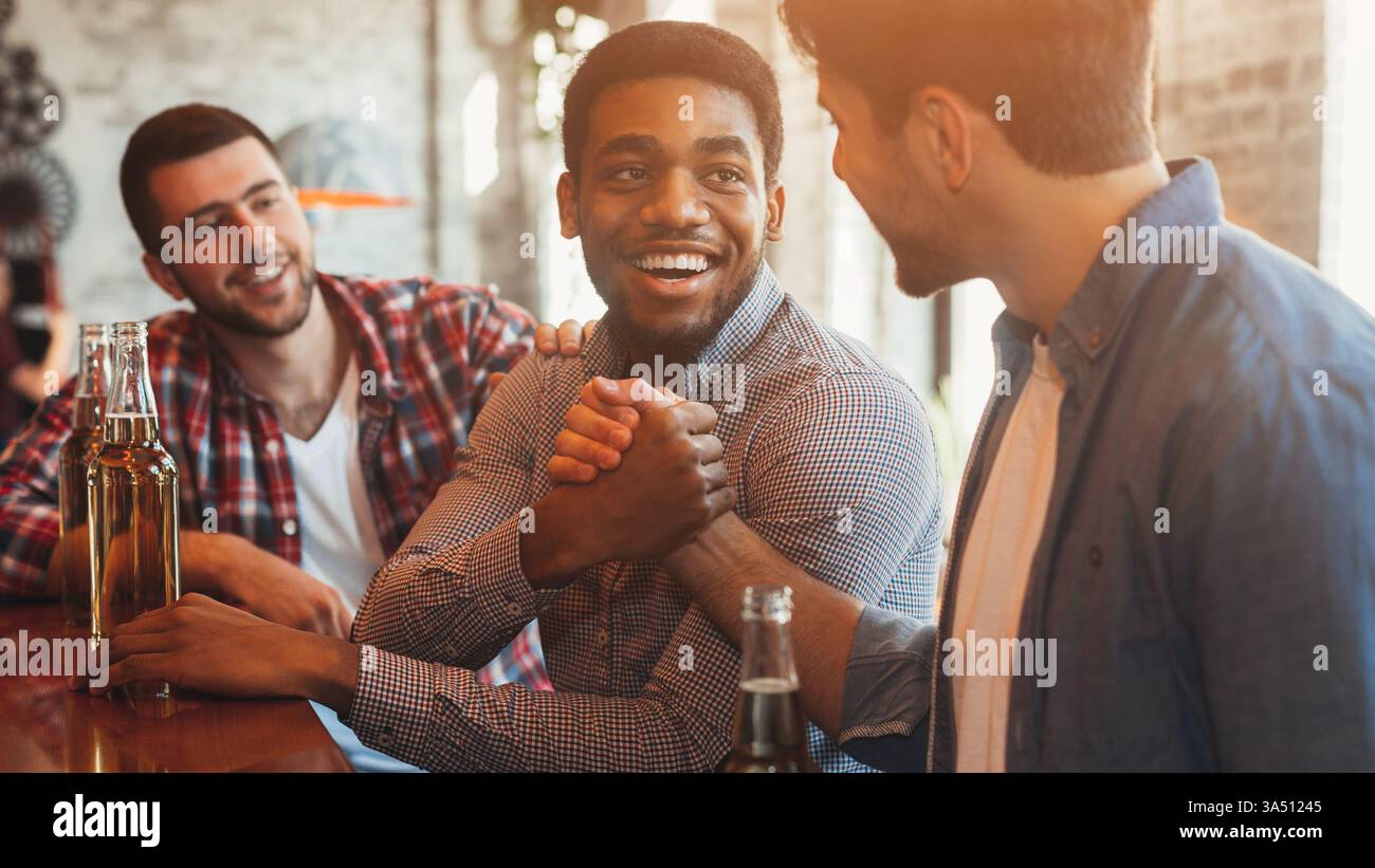 Les hommes saluent et serrent la main dans un pub, capturant un moment social détendu. Ils sourient et discutent autour d'un verre, véhiculant amitié et camaraderie. Utile pour le marketing, la socialisation et les visuels de réseautage sur le lieu de travail. Banque D'Images