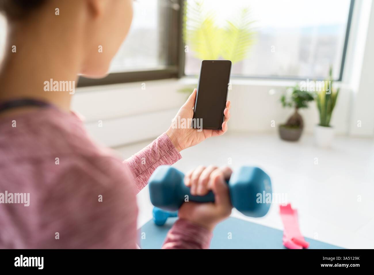 La femme chinoise asiatique dans les vêtements de fitness décontractés utilise un smartphone pour suivre une vidéo d'entraînement à la maison. La scène de fitness en intérieur propose des poids et un entraînement en ligne, adaptés aux campagnes de santé, de bien-être et de fitness numérique. Idéal pour les articles sur la perte de poids, les applications de fitness à domicile, ou le coaching virtuel. Banque D'Images
