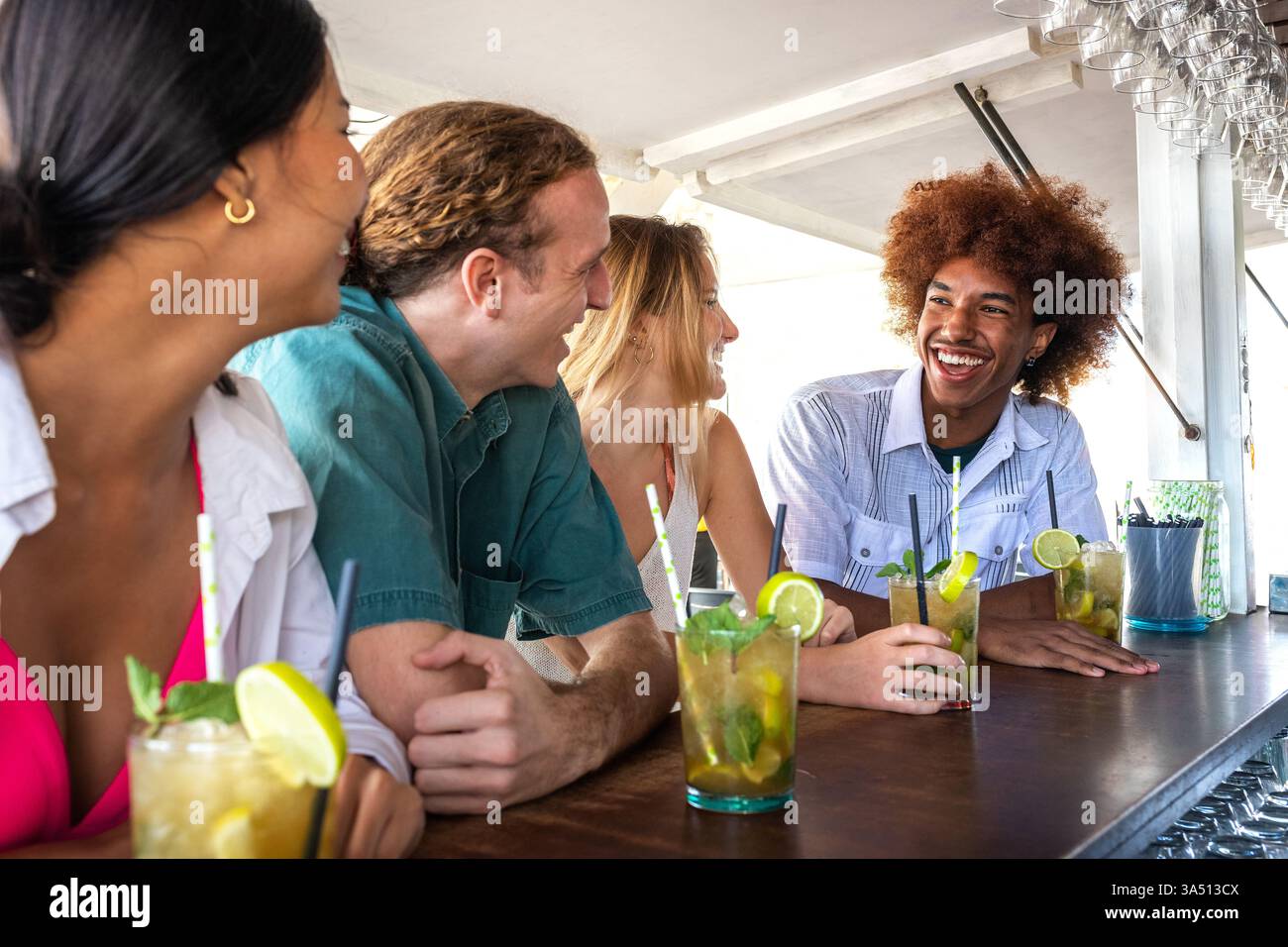 Groupe diversifié d'amis dans un bar de plage, bavarder et rire tout en dégustant des cocktails. Ils sont assis ensemble au comptoir sous le soleil d'été, capturant les vibrations de vacances insouciantes. Idéal pour le style de vie, l'amitié, les voyages et les scènes sociales. Banque D'Images