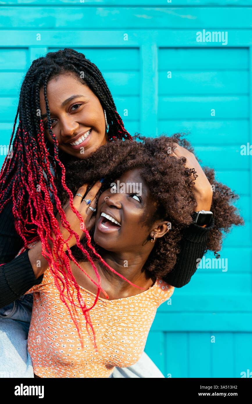 Femme noire donnant piggyback ride à une amie femelle contre le mur bleu à l'extérieur. Un moment d'amitié ludique parfait pour les campagnes lifestyle, sociales et de bien-être. Le sourire et l’énergie véhiculent chaleur et camaraderie. Banque D'Images