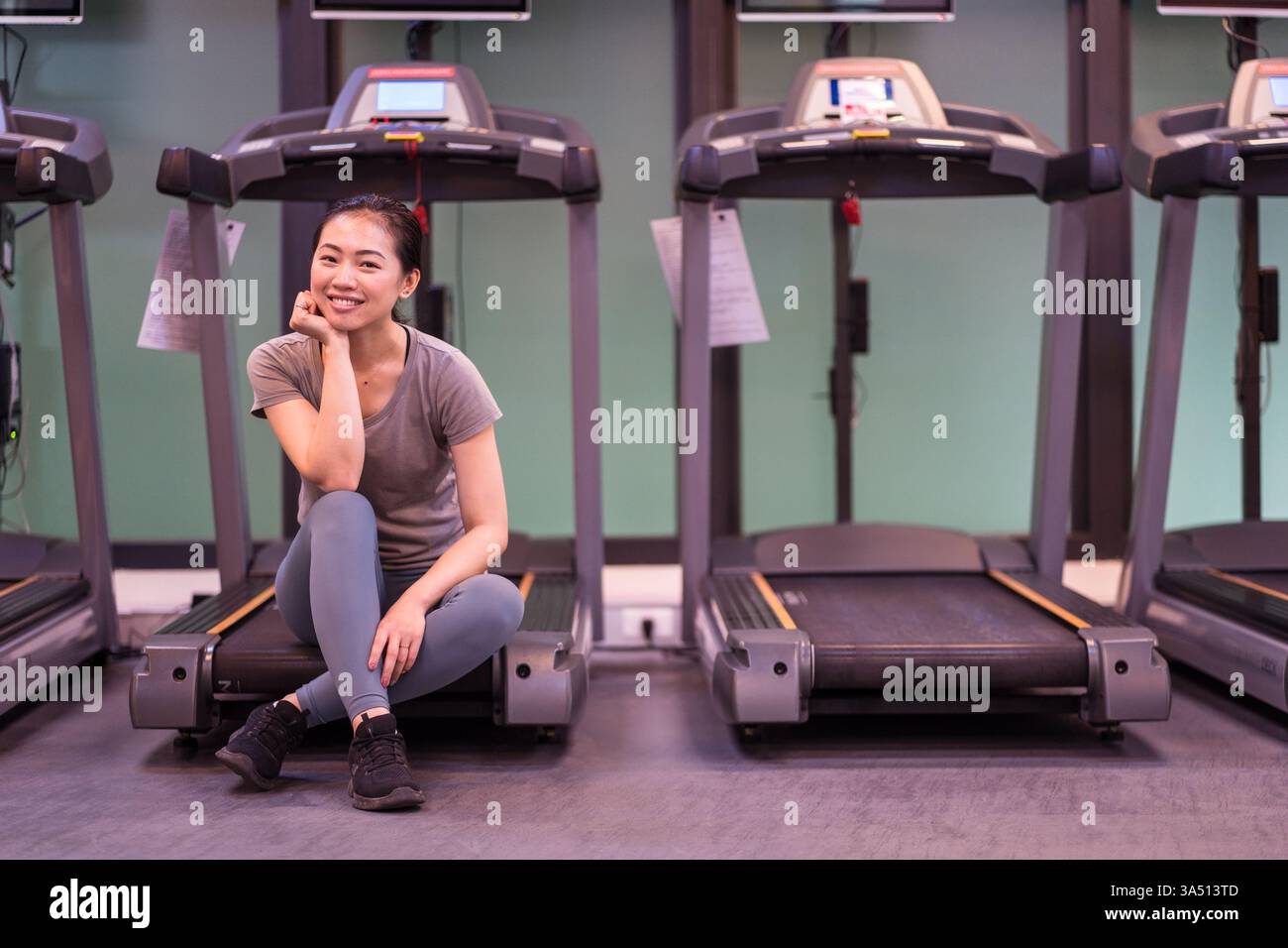 Athlète femme asiatique dans des vêtements d'activité élégants repose sur un tapis roulant après une séance d'entraînement dans une salle de sport moderne. Cette image met en valeur la forme physique, la récupération et l'entraînement en salle dans un cadre de gymnastique contemporain. Banque D'Images