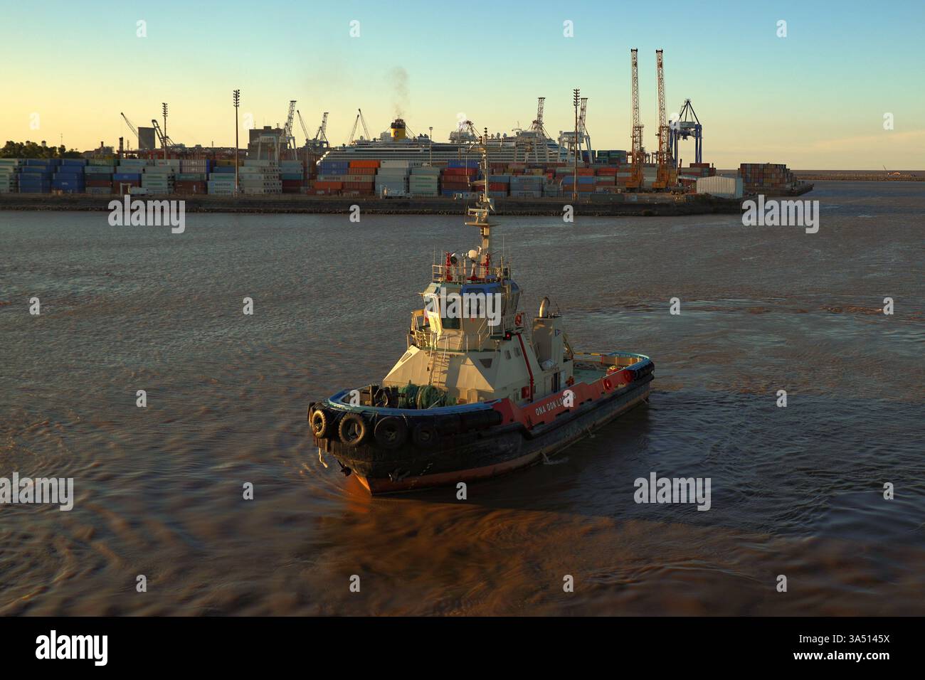 Remorqueur naviguant autour d'un quai de conteneurs au port de Buenos Aires, Argentine, avec un bateau de croisière au loin au coucher du soleil Banque D'Images