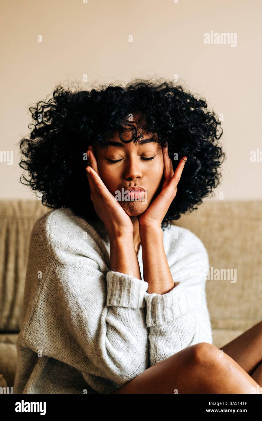 Une femme noire est assise à la maison, faisant un visage de canard ludique avec les yeux fermés. Le moment décontracté et attachant transmet la vie à la maison détendue et l'expression personnelle. Idéal pour le style de vie, les portraits de style médias sociaux et les scènes féminines de tous les jours. Banque D'Images