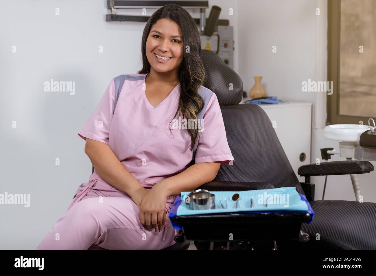 Dentiste hispanique souriante en uniforme rose est assise près d'un fauteuil dentaire dans une clinique. Une scène de soins de santé conviviale adaptée à la pratique dentaire, aux soins bucco-dentaires et aux sujets de soins aux patients. Des images professionnelles et accessibles. Banque D'Images