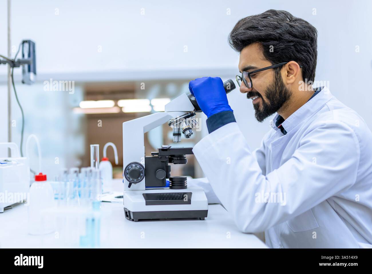 Un scientifique aux cheveux foncés mène des expériences dans un laboratoire moderne, ajustant un échantillon dans une boîte de Pétri tout en regardant dans un microscope. Une scène scientifique et biotechnologique mettant en vedette des équipements de laboratoire et des travaux de biologie moléculaire. Convient pour l'éducation, la recherche et les matériaux de communication scientifique. Banque D'Images