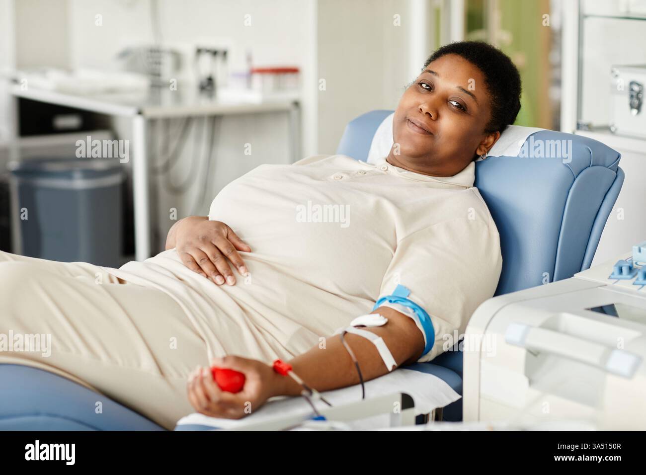 Portrait d'une femme souriante faisant don de sang dans un centre de don, assise sur une chaise. L'image communique la santé, la charité et la vie communautaire. Idéal pour les campagnes de don de sang, les soins de santé et les initiatives de sauvetage. Banque D'Images