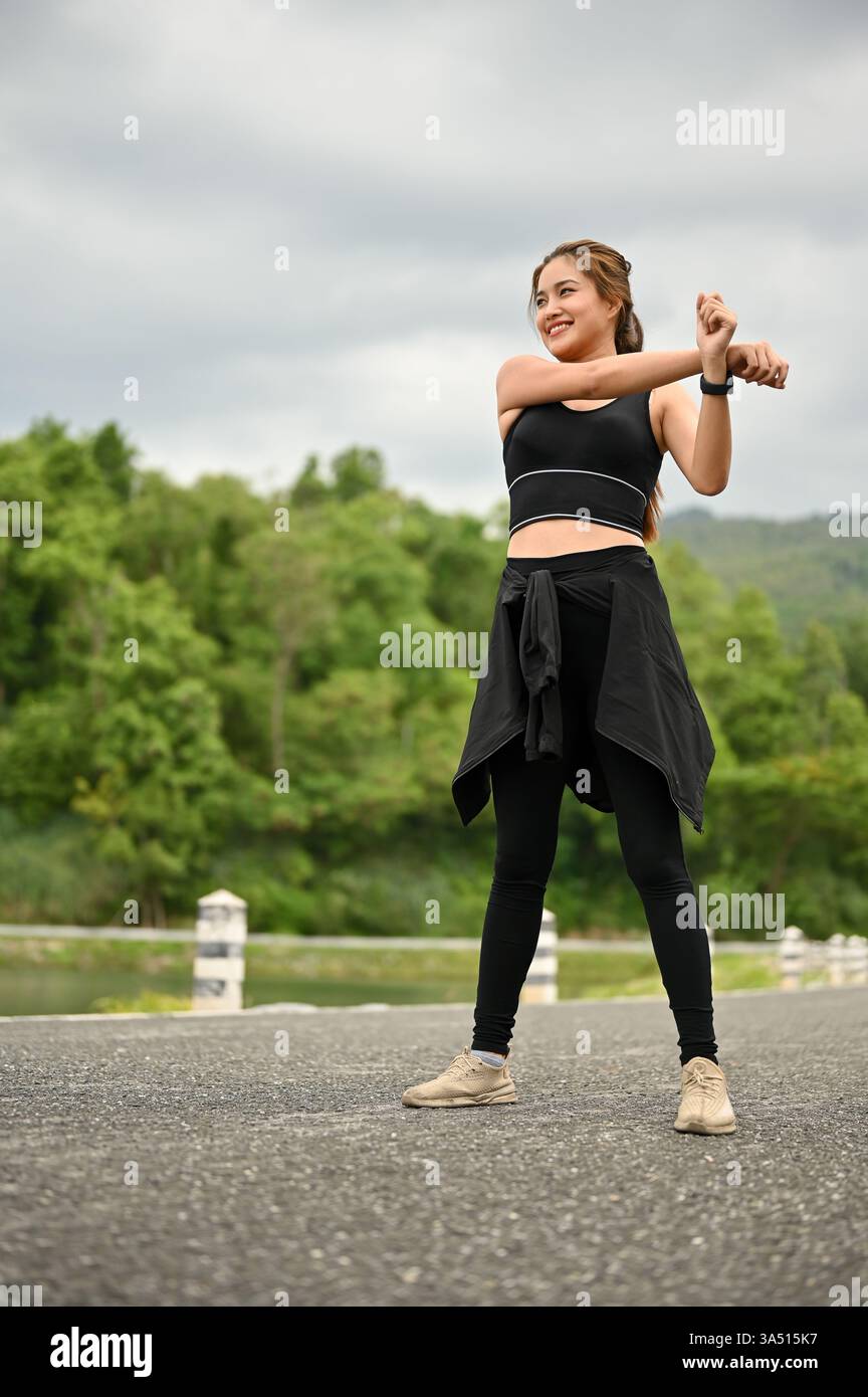 Portrait vibrant d'une jeune femme asiatique sportive en tenue de sport s'échauffant avant une course dans un parc public. Elle étire ses bras avec détermination, soulignant la forme physique et une vie saine en plein air. Idéal pour les vêtements de sport, le bien-être et les projets de style de vie actif. Banque D'Images
