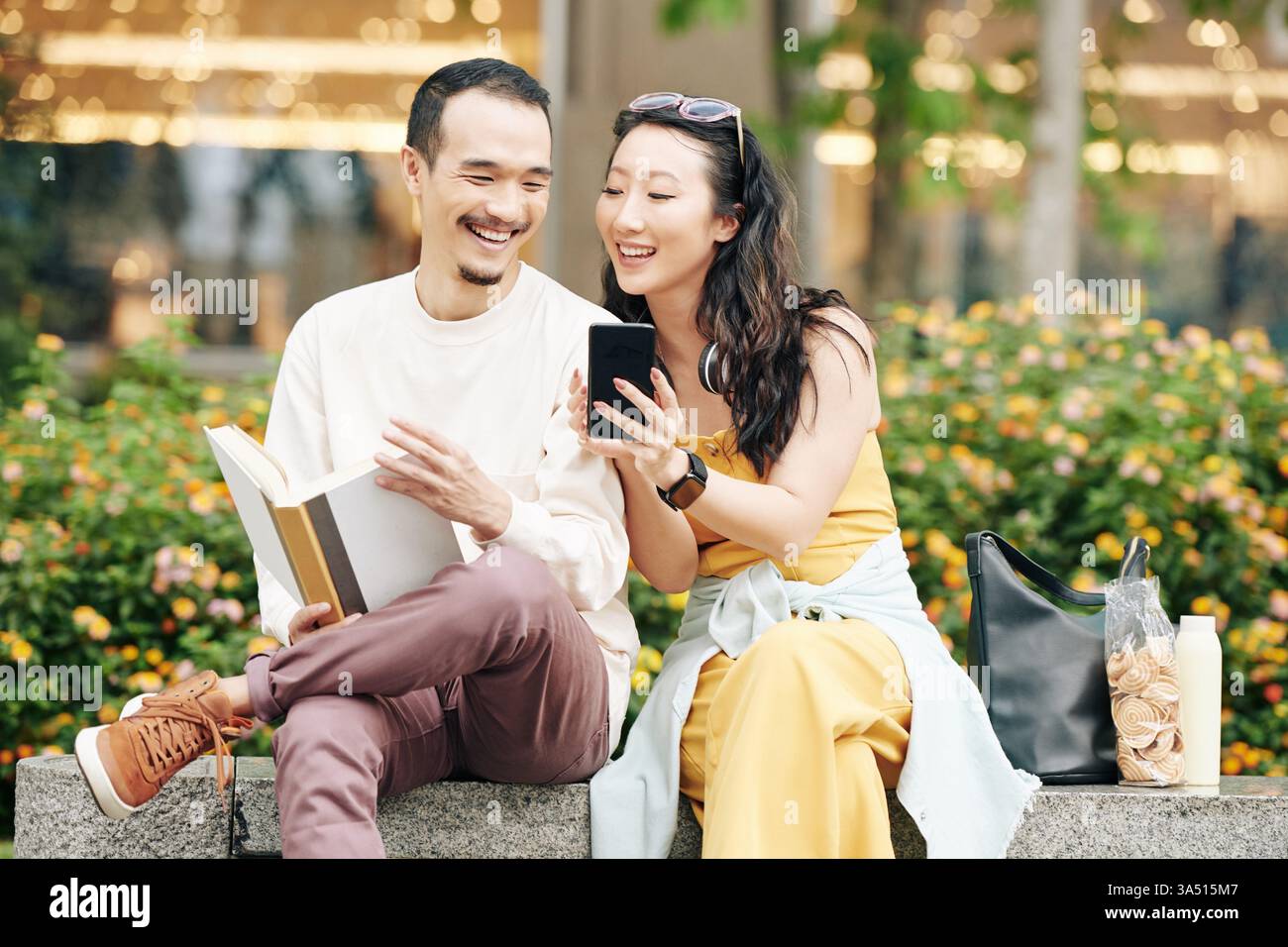Heureux jeune couple asiatique riant à un mème sur un smartphone tout en étant assis sur un banc de parc avec des collations. Un cadre extérieur décontracté et une interaction chaleureuse le rendent idéal pour les campagnes de rencontres, de style de vie ou de médias sociaux. Parfait pour le contenu sur la convivialité, les loisirs et la technologie. Banque D'Images