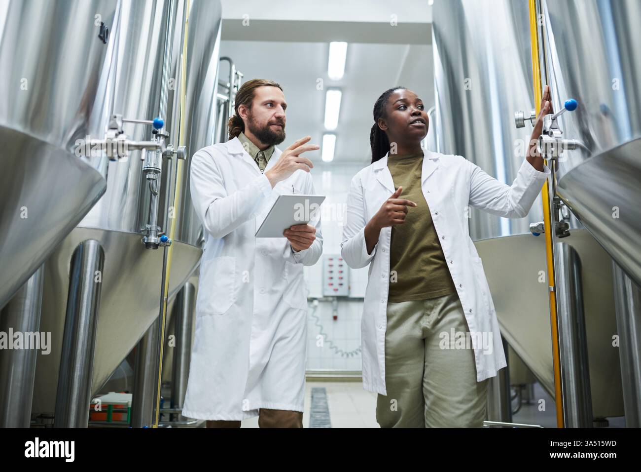 Un groupe diversifié de technologues de brasserie inspecte l'équipement dans un cadre d'usine. Ils portent des blouses de laboratoire et utilisent une tablette pour surveiller la production et assurer la qualité. Banque D'Images
