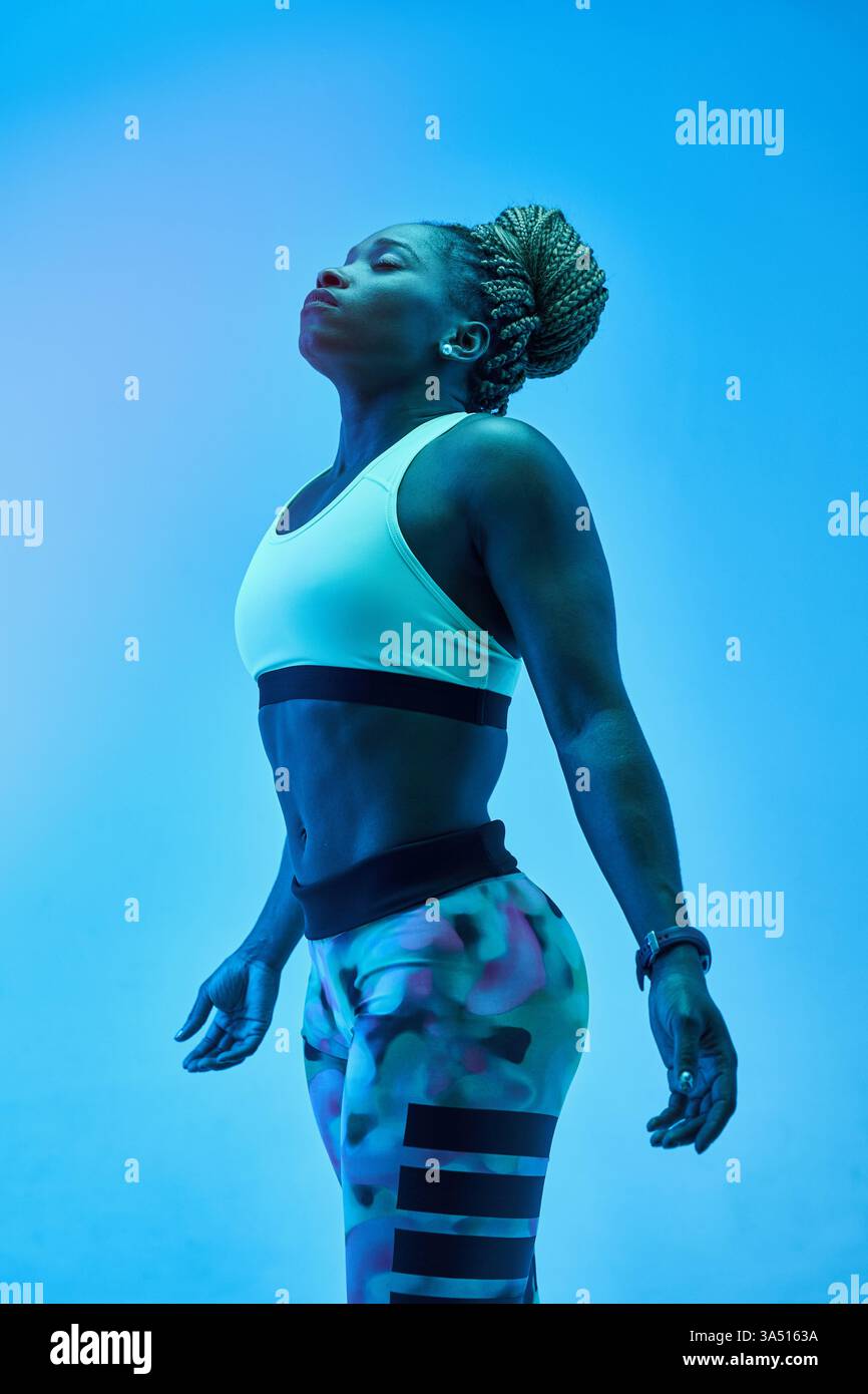 Athlète féminine noire se tient debout avec la tête inclinée vers l'arrière et les yeux fermés, sur un fond de studio bleu. Cette image transmet l'énergie, la concentration et la pleine conscience dans des contextes de fitness et de bien-être. Idéal pour les vêtements de sport, l'entraînement et les campagnes de style de vie actif. Banque D'Images