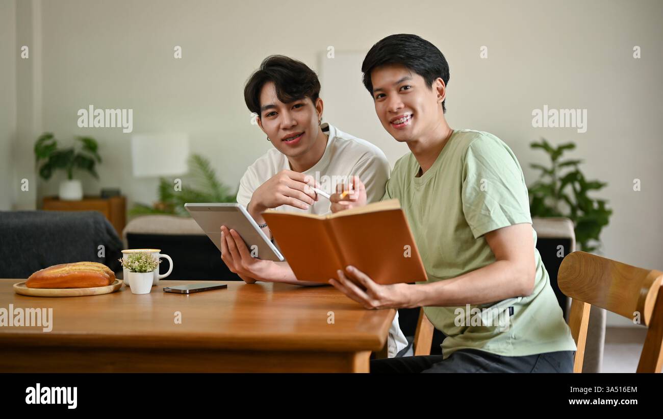 Souriants étudiants asiatiques étudiant ensemble à la maison, tenant un livre et une tablette à une table. Cette scène de style de vie éducatif souligne l'amitié, l'apprentissage et l'étude à distance, idéal pour les caractéristiques de la vie universitaire. Banque D'Images