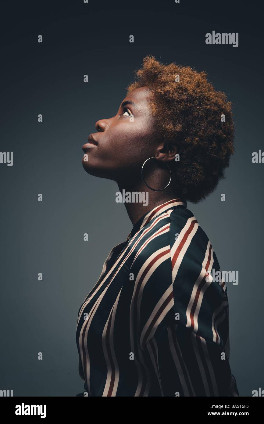 Femme noire sérieuse avec un Afro se tient sur le côté dans un studio sombre, créant un portrait sombre. Ce cliché studio met l'accent sur la confiance, l'individualité et le style contemporain. Banque D'Images