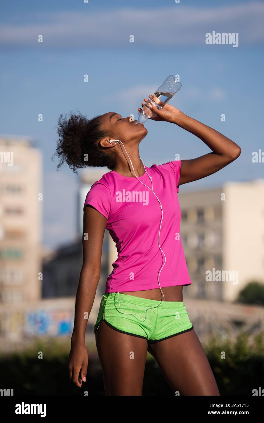 Une jeune femme afro-américaine en tenue athlétique fait une pause après un jogging en ville, buvant de l'eau avec des écouteurs. Elle respire le fitness et la santé urbaine, parfait pour la santé, le bien-être et le mode de vie actif. Banque D'Images