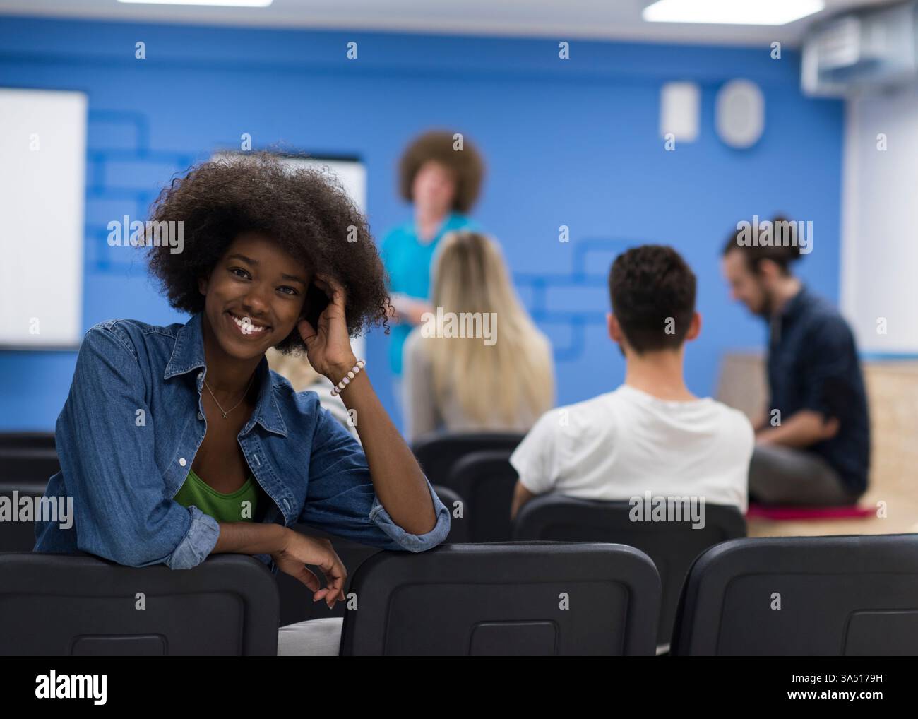 Joyeuse femme noire avec un afro est assise en arrière dans une chaise, s'engageant avec des collègues dans une salle de conférence. L'ambiance moderne du lieu de travail met en valeur la diversité, le travail d'équipe et la créativité. Idéal pour les affaires, la culture du lieu de travail et les thèmes du travail d'équipe. Banque D'Images