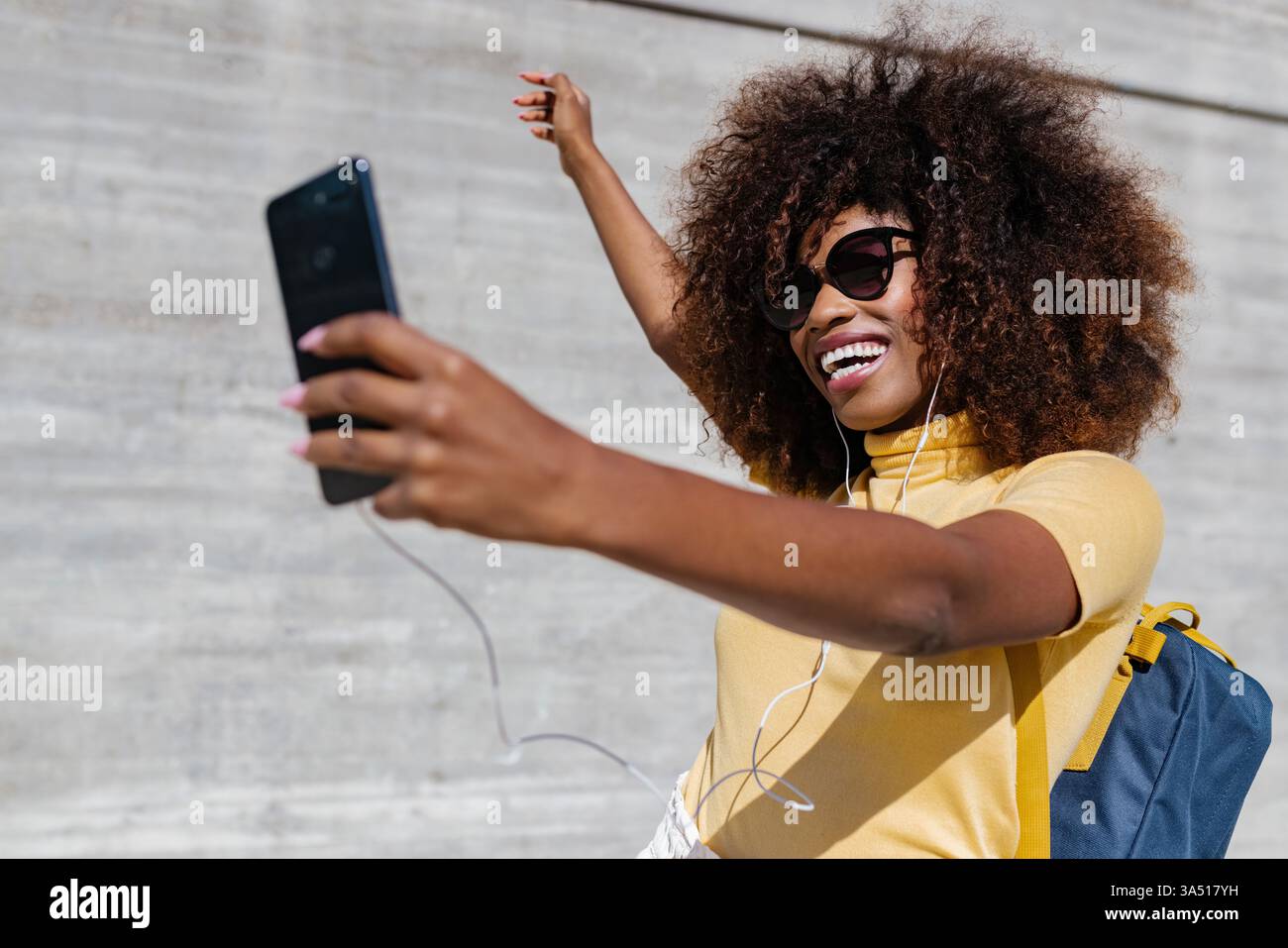 Joyeuse femme afro-américaine prend un selfie avec un smartphone contre un mur gris. Image de style de vie urbain décontracté adapté aux médias sociaux, à l'image de marque personnelle et à la vie moderne. Parfait pour le bien-être, l'autonomisation et les campagnes de style de vie des jeunes. Banque D'Images