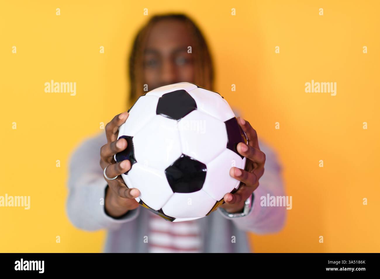 Portrait studio d'un homme noir dans une chemise rayée rouge et une veste tenant un ballon de football sur un fond jaune vif. Un concept sportif dynamique adapté au style de vie sportif et aux images de sports d'équipe. Banque D'Images