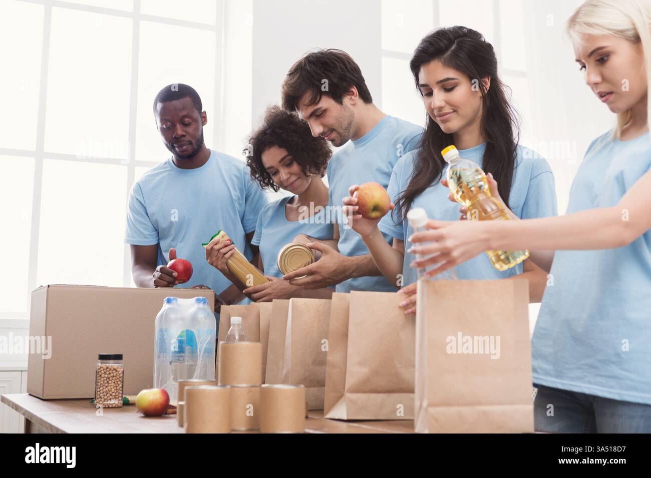 Divers bénévoles en chemises bleues emballant les dons alimentaires dans des sacs en papier à l'intérieur. L'équipe souriante fait preuve de travail d'équipe et de charité en action. Idéal pour les visuels de charité, de bénévolat et de soutien communautaire. Banque D'Images