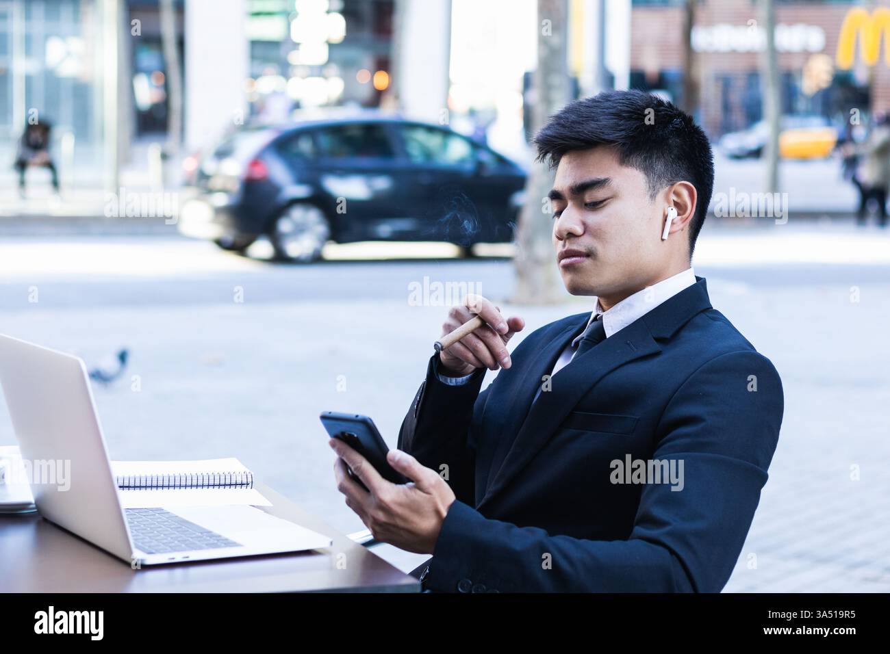 Homme d'affaires asiatique sérieux portant des écouteurs sans fil est assis dans un café en plein air avec un ordinateur portable, à l'aide d'un smartphone et tenant un cigare. Cette scène d’affaires contemporaine transmet la connectivité, l’entrepreneuriat et le travail ciblé. Banque D'Images
