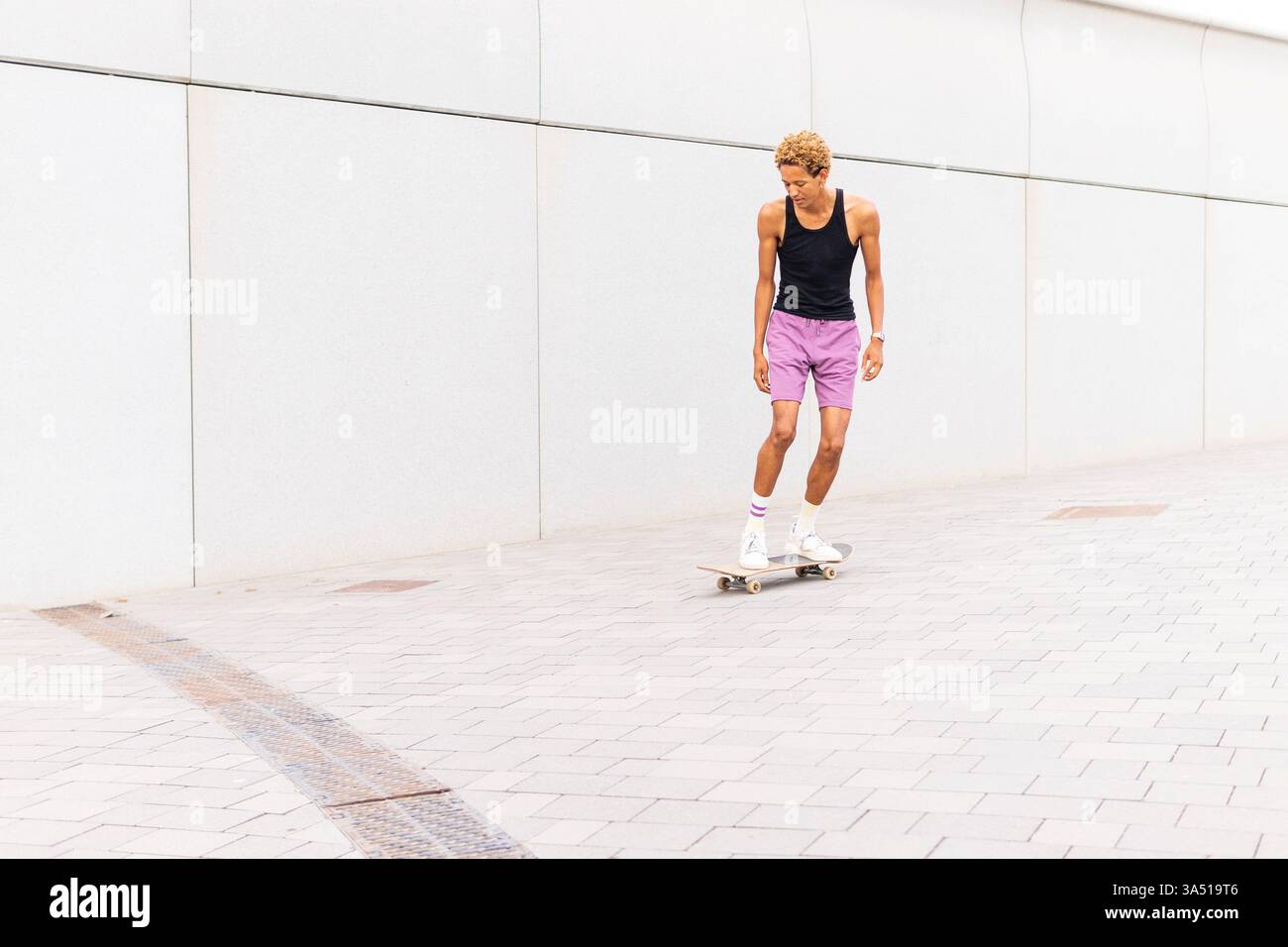 Photo du corps entier d'un jeune Afro-américain patinant sur une passerelle pavée de la ville. Il s'entraîne dans la rue avec une énergie dynamique dans des vêtements sportifs décontractés. Parfait pour le sport urbain, le style de vie urbain et les visuels de fitness. Banque D'Images