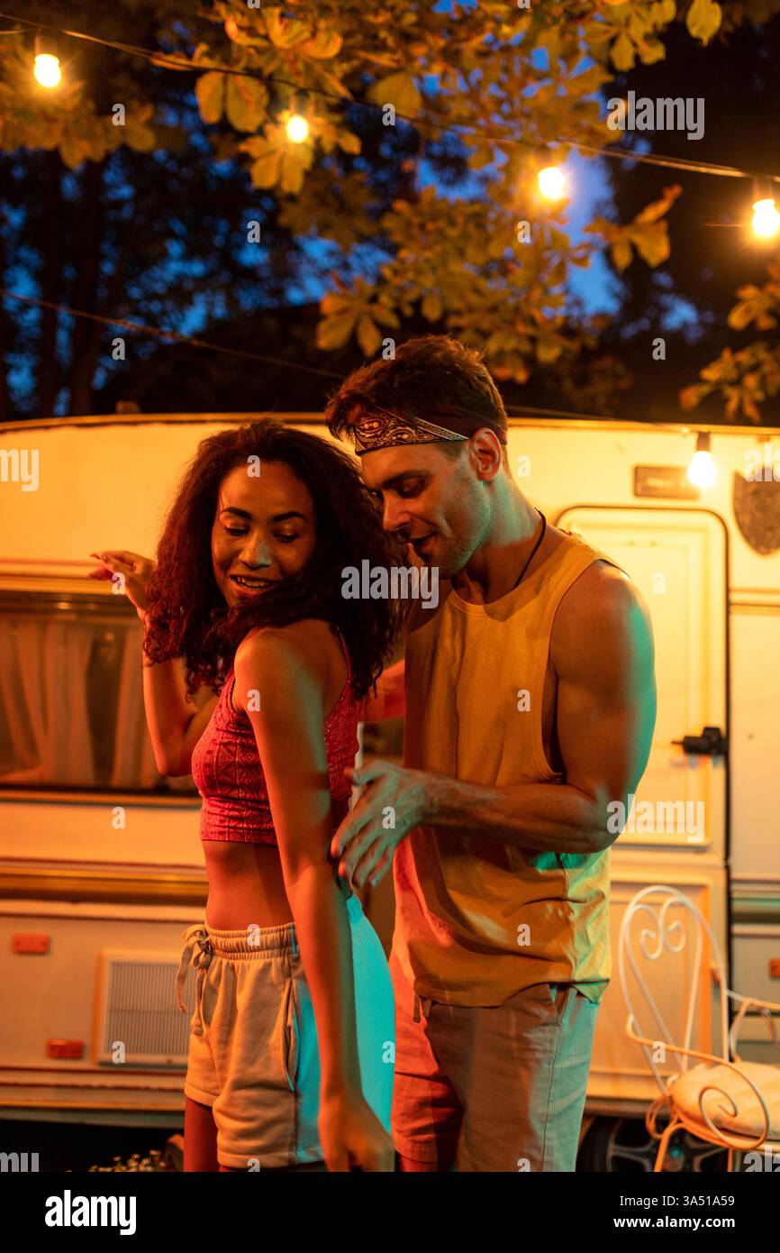 Un jeune couple dansant à côté d'un camping-car la nuit pendant des vacances. La scène capture la soif de vagabondage, les voyages en voiture et le style de vie des voyages en plein air. Parfait pour les voyages, le camping et les escapades romantiques. Banque D'Images