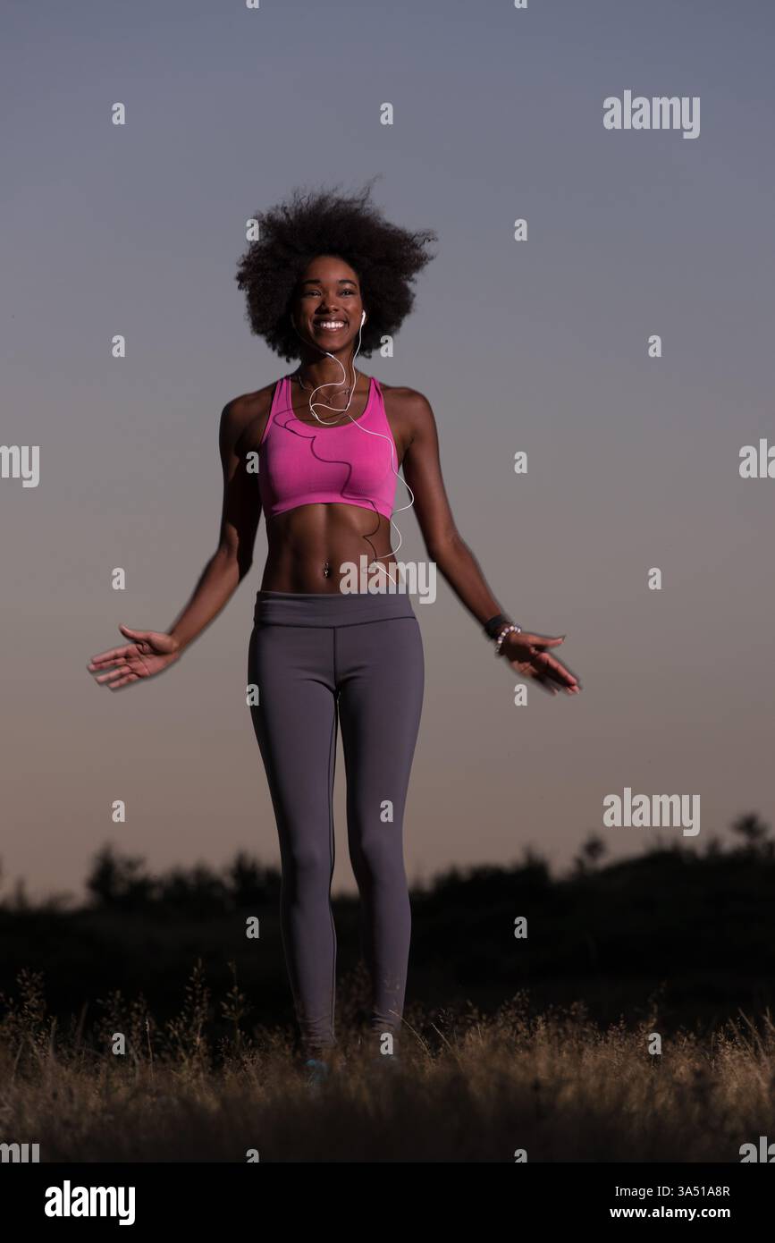 Jeune femme noire en bonne santé faisant des exercices d'étirement dans une prairie au coucher du soleil d'été. Elle se réchauffe après le jogging, capturant un moment de fitness actif en plein air. Banque D'Images