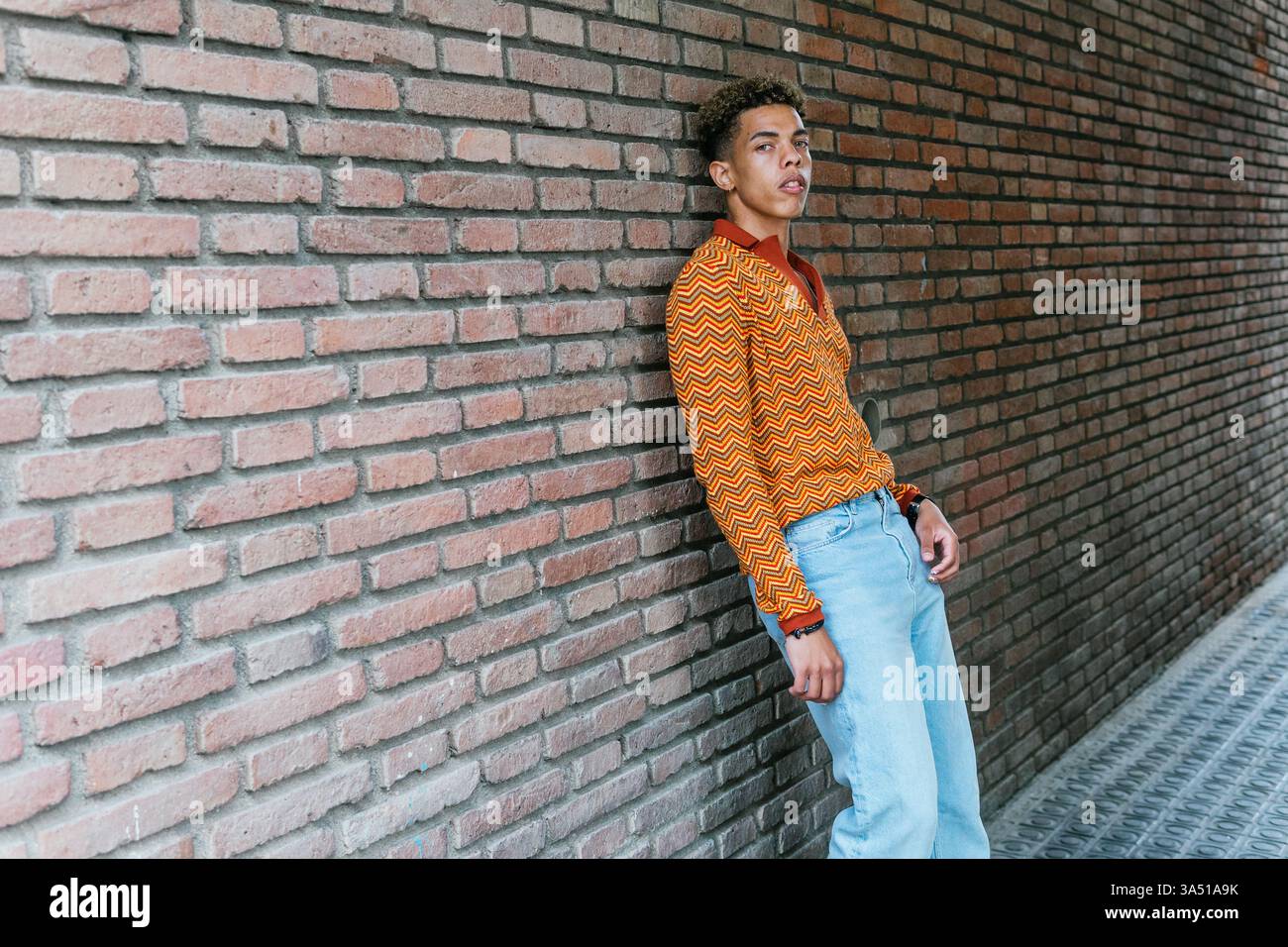 Homme hispanique sérieux s'appuie contre un mur de briques dans une rue de la ville, projetant une ambiance urbaine confiante et élégante. Le portrait moderne fonctionne bien pour la mode, le style de rue et le marketing masculin contemporain. Une image polyvalente pour les campagnes de style de vie urbain. Banque D'Images