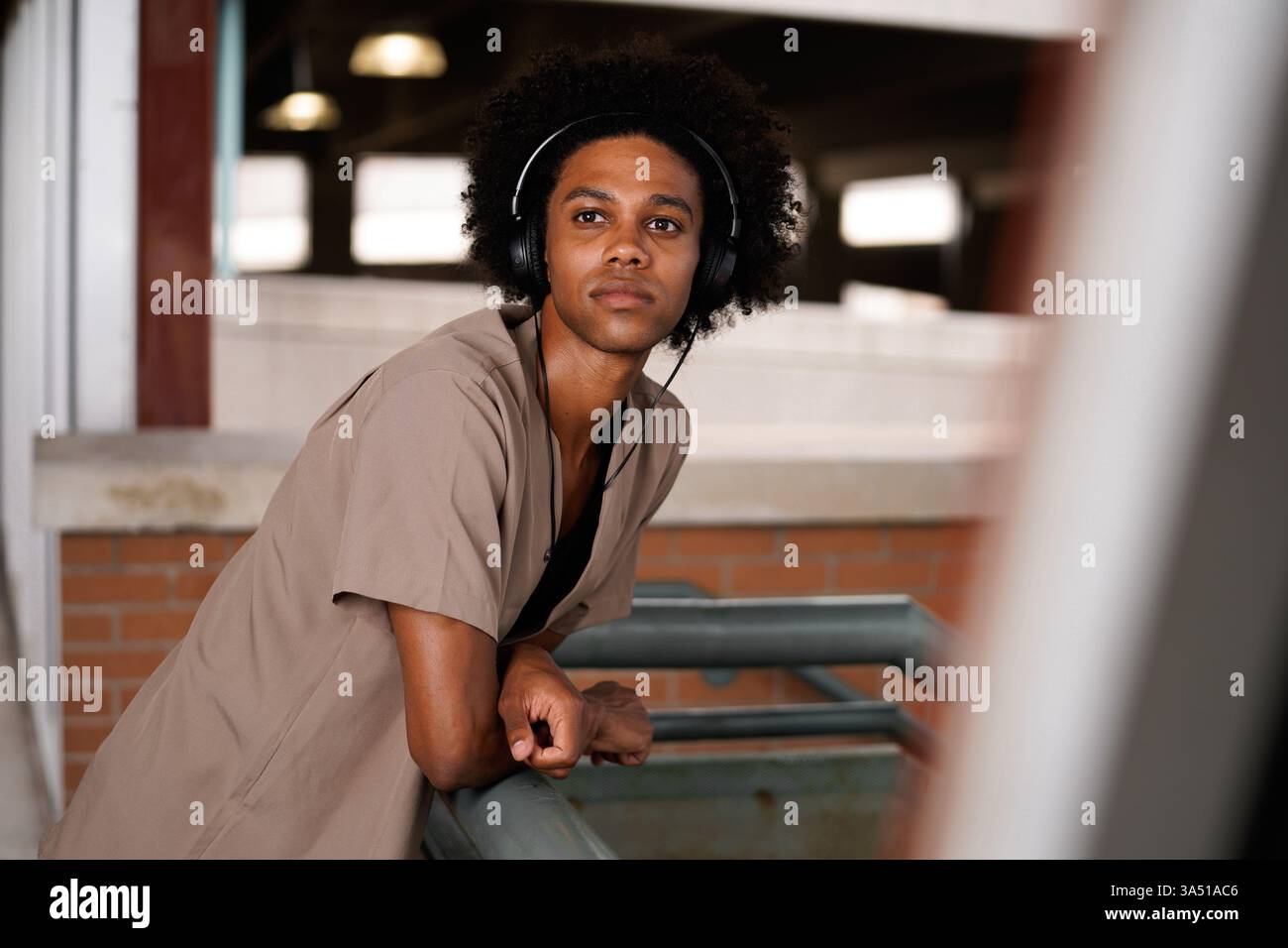 Homme noir avec Afro bouclé portant des écouteurs écoute de la musique tout en se penchant sur une balustrade à une gare. Une image de style de vie urbain et contemporain parfaite pour la musique, les déplacements et les campagnes de vie urbaine. Idéal pour exprimer votre style personnel et votre concentration. Banque D'Images