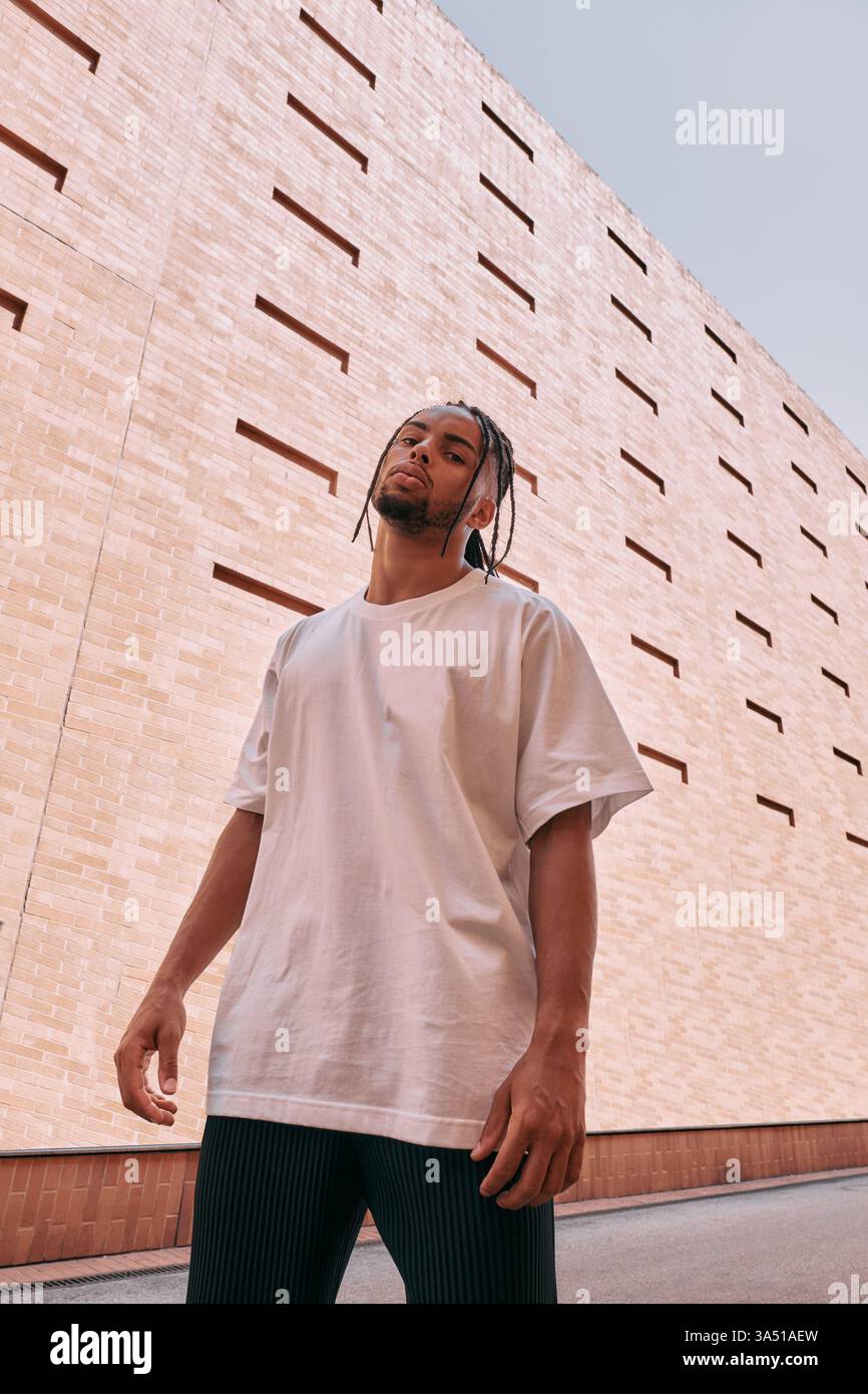 Homme afro-américain debout dehors près d'un bâtiment, regardant vers le bas avec une pose réfléchie. Image de style de vie urbain adaptée à la mode, au style de rue ou aux thèmes de développement personnel. Portrait clair et moderne en lumière naturelle. Banque D'Images