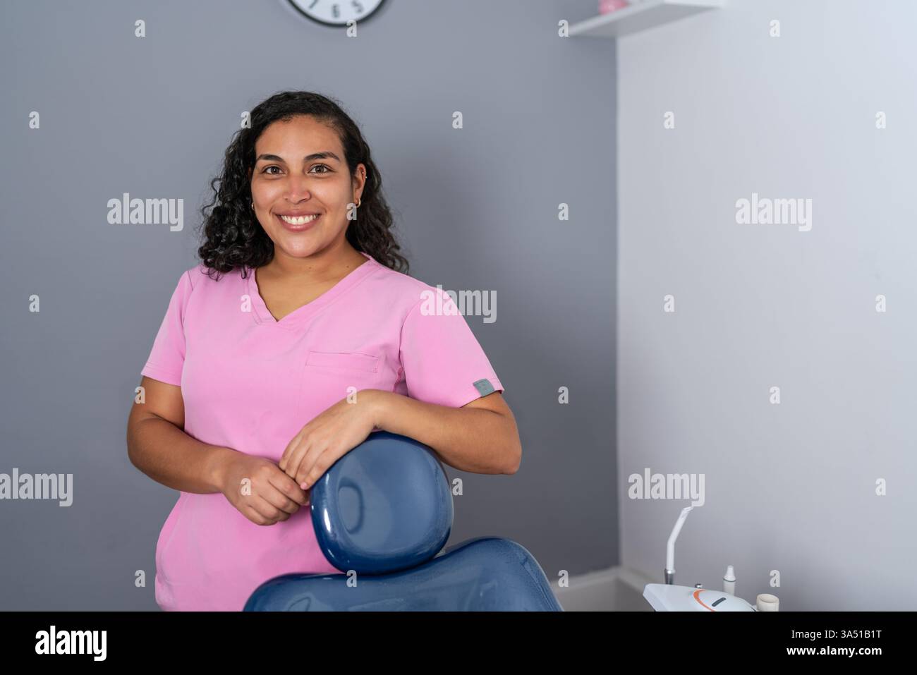 Dentiste hispanique souriante avec les cheveux bouclés appuyés sur une chaise dentaire dans une clinique moderne. Idéal pour les soins dentaires, la santé bucco-dentaire et les visuels professionnels de la santé. Banque D'Images