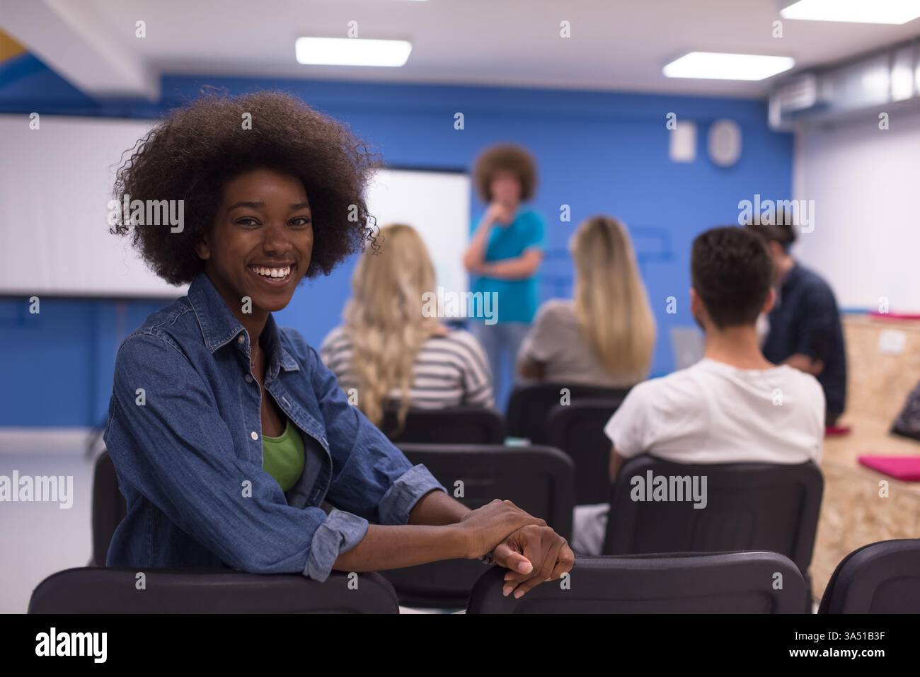 Joyeuse femme noire avec un afro s'assoit à l'arrière sur une chaise à côté de collègues dans une salle de conférence. Cette image moderne du lieu de travail convient au contenu sur le travail d'équipe, le leadership et les environnements d'affaires diversifiés. Banque D'Images