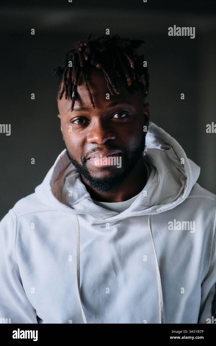 Jeune homme afro-américain dans un sweat à capuche sourit à la caméra dans un cadre intérieur sombre. L'arrière-plan sombre avec éclairage intérieur souligne son ambiance confiante et accessible, adaptée aux campagnes de mode et de style de vie. Idéal pour les publicités mettant en vedette le streetwear décontracté, le style urbain jeunesse, ou l'image de marque personnelle. Banque D'Images