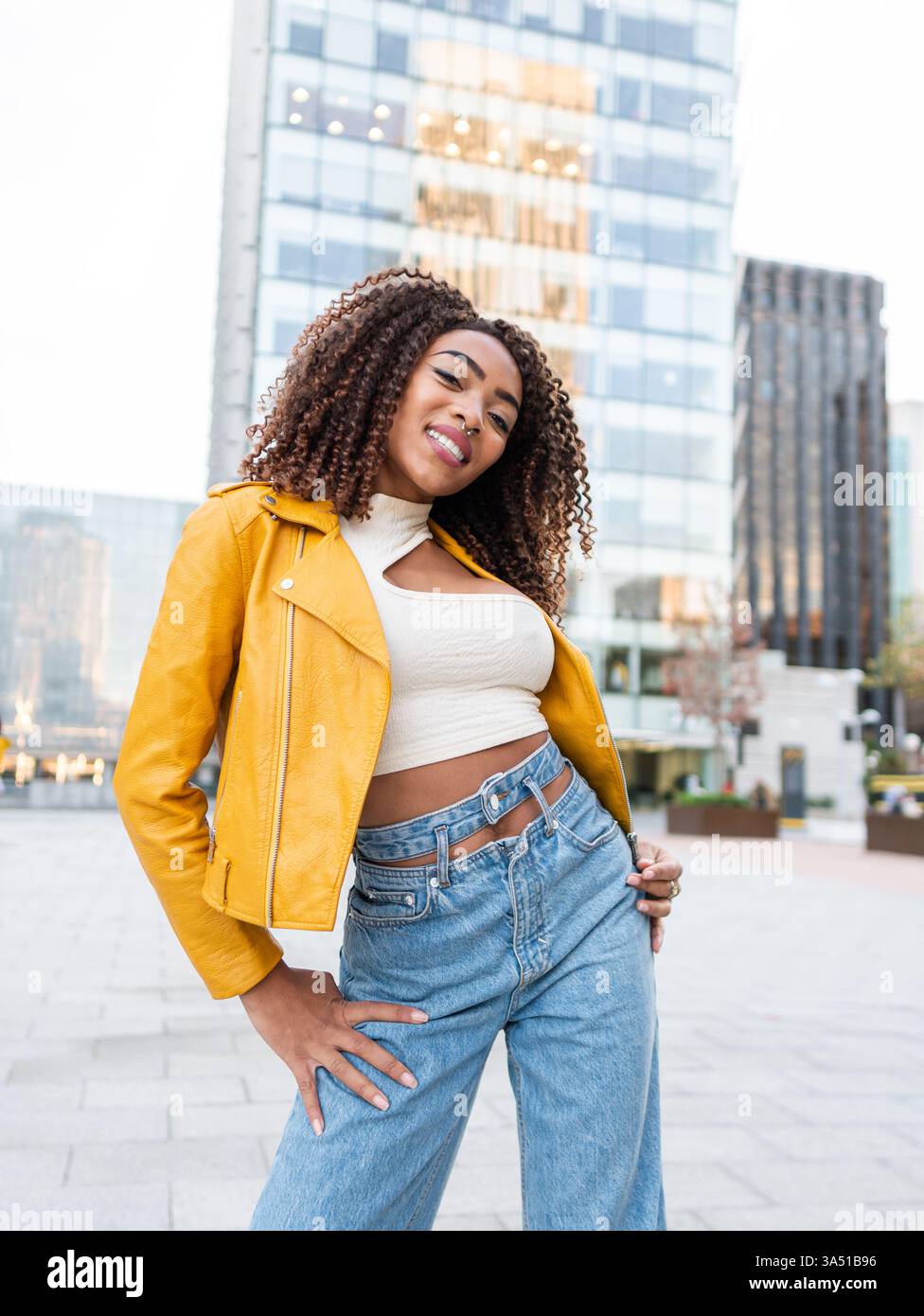 Femme hispanique souriante dans une veste en cuir jaune se tient dans une rue de la ville pendant la journée. Son style de rue et son expression ensoleillée véhiculent la mode urbaine et les vibrations positives. Parfait pour les campagnes de mode, de portrait et de style de vie urbain. Banque D'Images