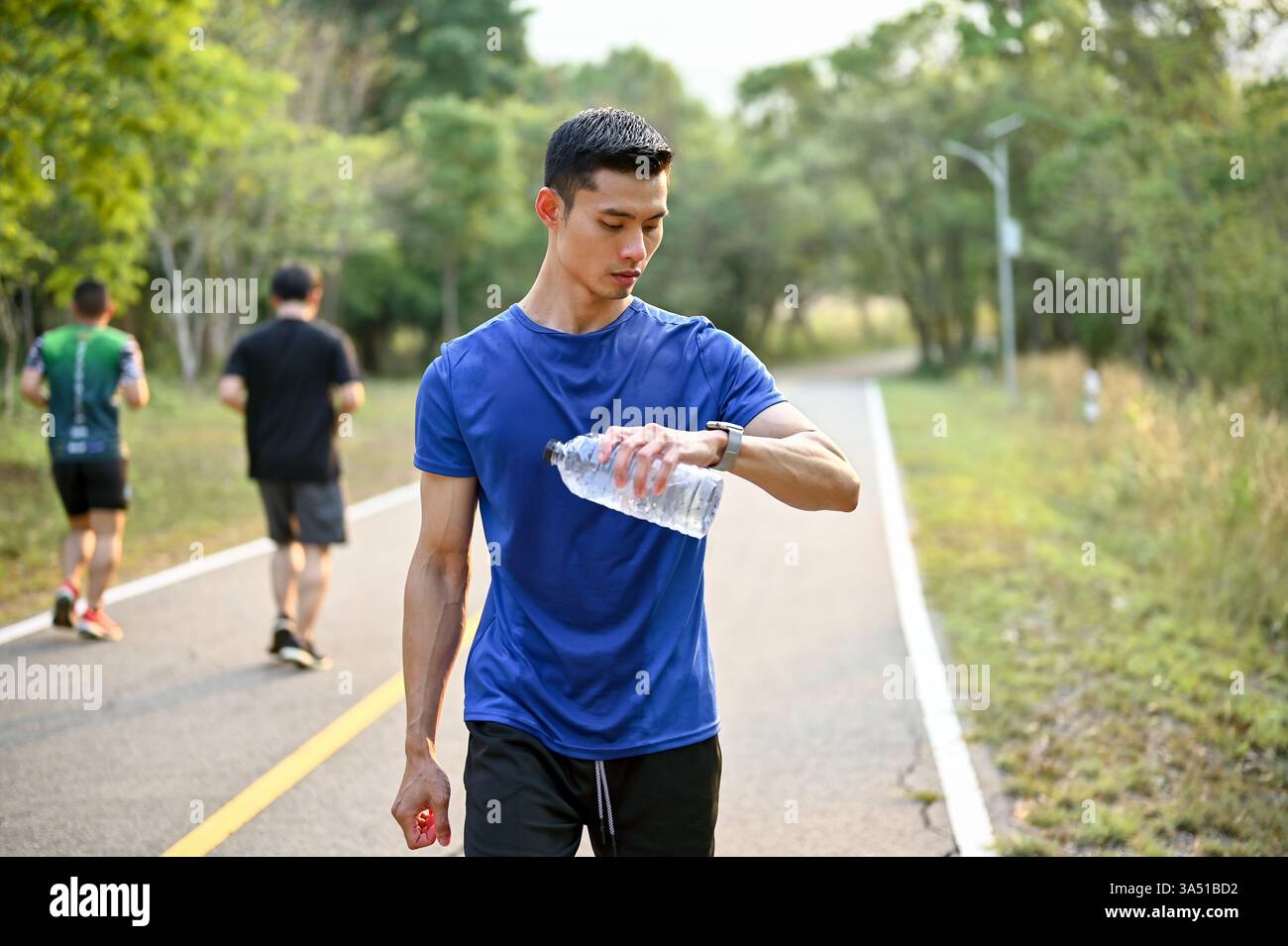 Homme asiatique millénaire en tenue athlétique vérifie sa fréquence cardiaque sur une smartwatch tout en faisant du jogging dans un parc. Idéal pour les campagnes de fitness, wearables et de style de vie actif. Banque D'Images