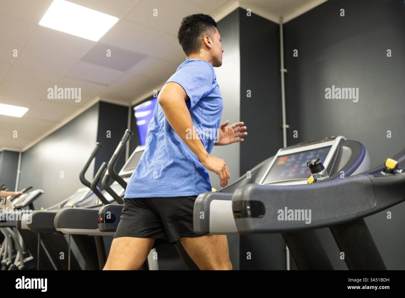 Jeune homme hispanique en vêtements de sport bleus court sur un tapis roulant dans une salle de sport moderne, en se concentrant sur le cardio. Cette scène de remise en forme intérieure met en évidence l'entraînement sportif, l'endurance et la santé générale. Banque D'Images
