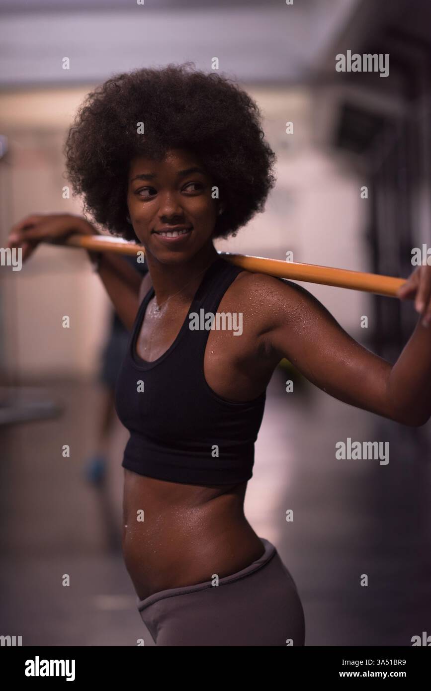 Jeune femme afro-américaine en vêtements de fitness se tient dans une salle de gym et soulève un bar vide avec un sourire. La scène met l'accent sur l'entraînement en gymnastique, la force et un style de vie actif. Banque D'Images