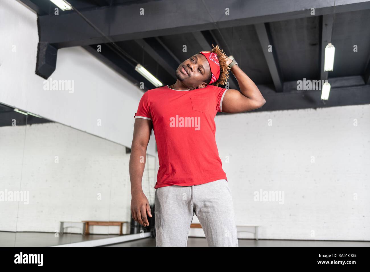 Homme noir en tenue sportive s'étire dans une salle de gym lumineuse, regardant loin de la caméra. Cette image convient aux campagnes de fitness et de santé, mettant en valeur l'énergie, la flexibilité et la concentration dans un environnement d'entraînement contemporain. Idéal pour les promotions de vêtements de sport et de studio. Banque D'Images