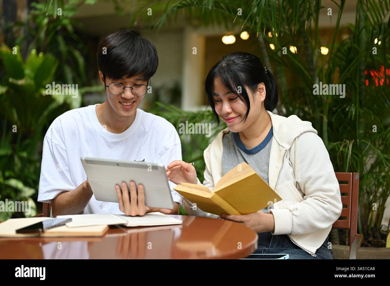 Deux étudiants asiatiques heureux assis dans un café en plein air, travaillant sur un projet scolaire ensemble. Ils discutent et partagent des idées dans un cadre lumineux, collégial-ville baigné de lumière naturelle. Idéal pour l'éducation, le travail d'équipe et les campagnes de style de vie des étudiants. Banque D'Images