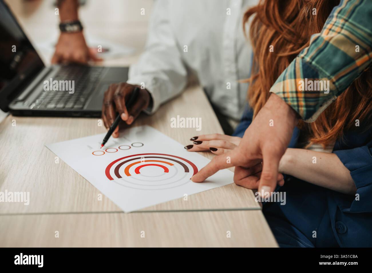 Une équipe diversifiée de collègues collabore sur un projet autour d'un bureau, avec un ordinateur portable et des graphiques. Les mains pointent vers les données, soulignant le travail d'équipe, la stratégie et un environnement de bureau moderne. Banque D'Images