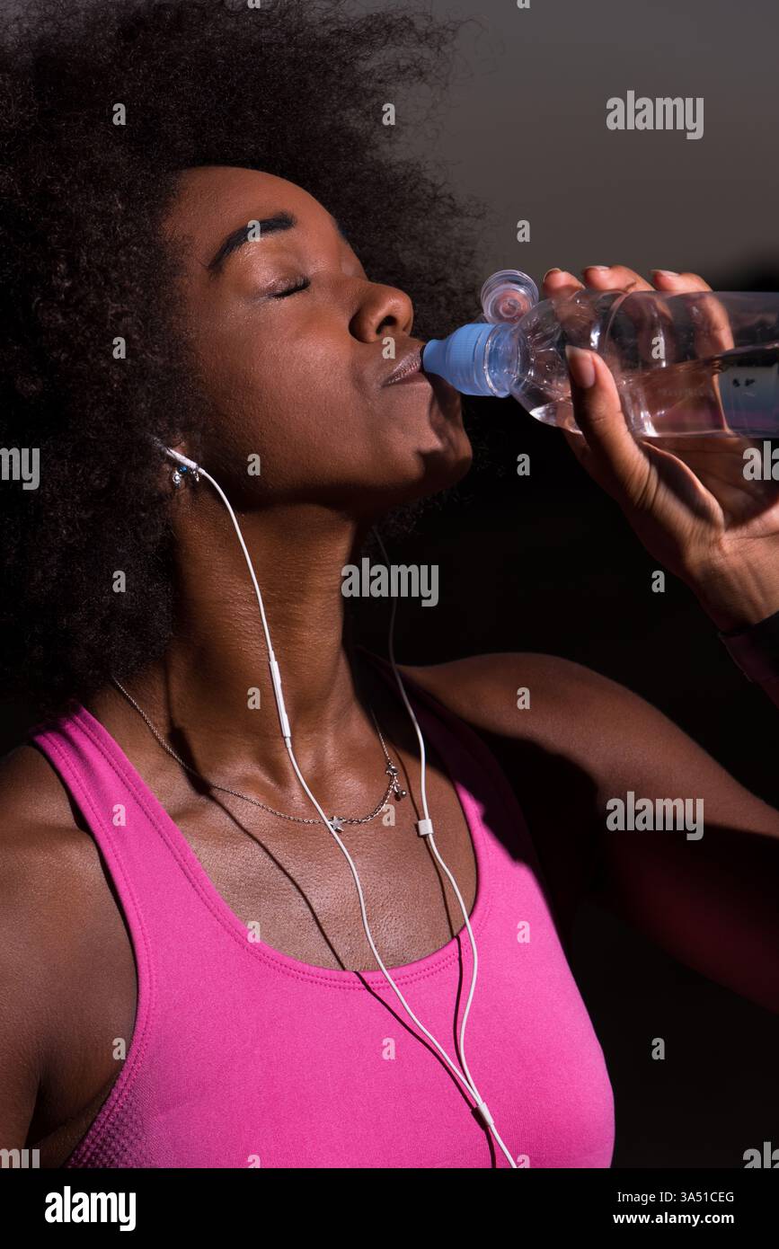 Jeune femme afro-américaine avec des écouteurs boire de l'eau après le jogging dans la nature sur une belle nuit d'été. La scène mêle fitness et vie en plein air avec une ambiance détendue. Idéal pour les campagnes de bien-être, de course à pied et de style de vie actif. Banque D'Images