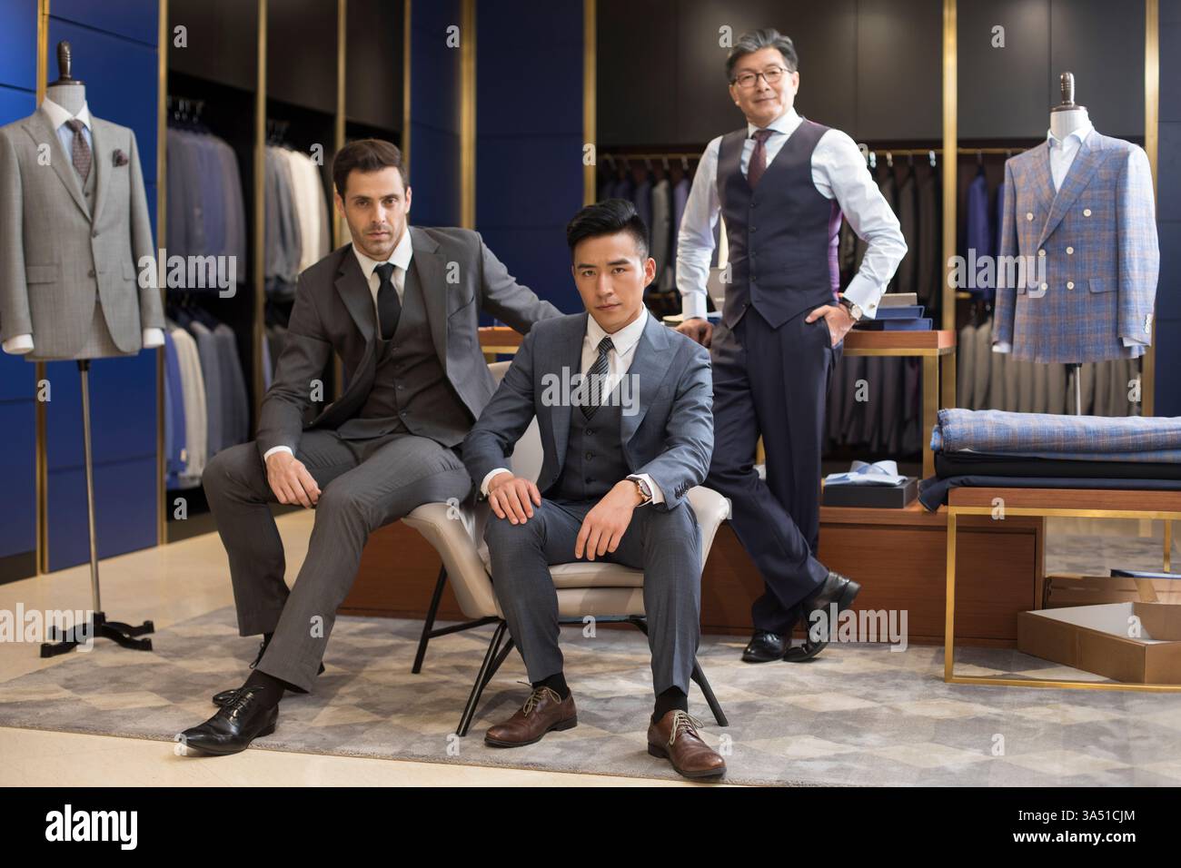 Un créateur de mode chinois se tient la main dans sa poche aux côtés d'un groupe diversifié d'hommes d'affaires chinois et hispaniques assis ensemble dans un studio de design. L'image exprime le leadership, l'expertise en design de mode et la collaboration dans un environnement moderne de vente au détail ou de studio. Idéal pour le contenu sur l'industrie de la mode, les vêtements de créateur et le travail d'équipe professionnel. Banque D'Images