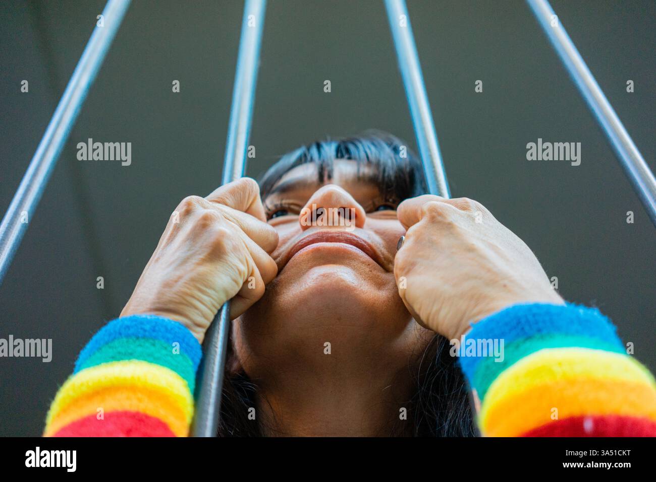 Femme hispanique portant un bracelet arc-en-ciel se tient derrière des garde-corps en métal à la maison, suggérant la résilience et la fierté quotidiennes. La scène mêle style personnel avec un sentiment de communauté et de liberté dans un cadre domestique. Idéal pour le marketing inclusif LGBTQ, les campagnes sociales ou le contenu de style de vie à la maison. Banque D'Images