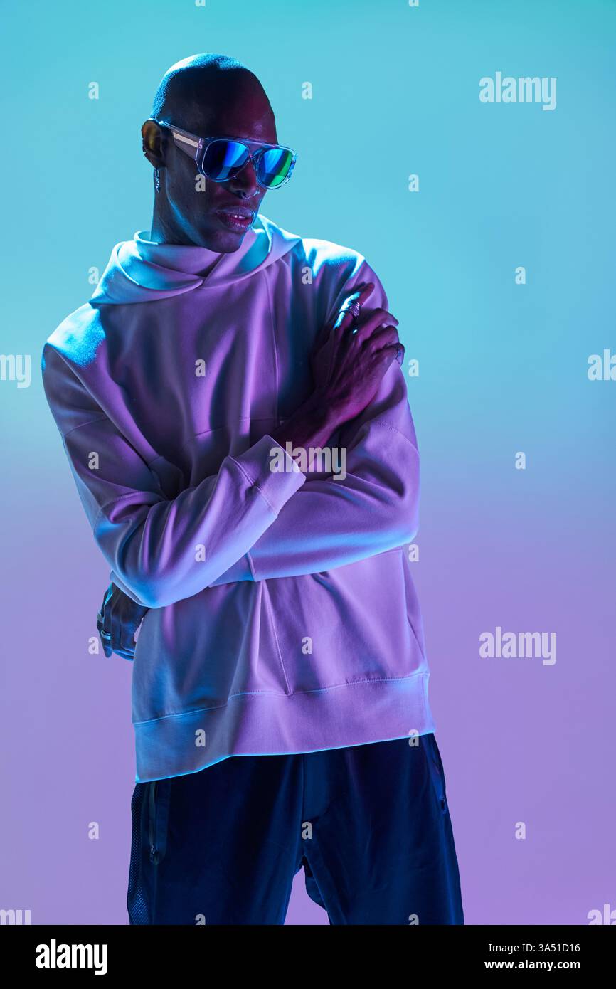 Vue de côté d'un homme afro-américain sérieux avec une tête rasée, portant une boucle d'oreille et des lunettes de soleil, posé dans un studio moderne avec un éclairage dramatique. Le look confiant et avant-gardiste convient aux vêtements pour hommes, au style et aux campagnes de marque. Idéal pour les éditoriaux de mode, les images de portfolio et l'esthétique urbaine contemporaine. Banque D'Images
